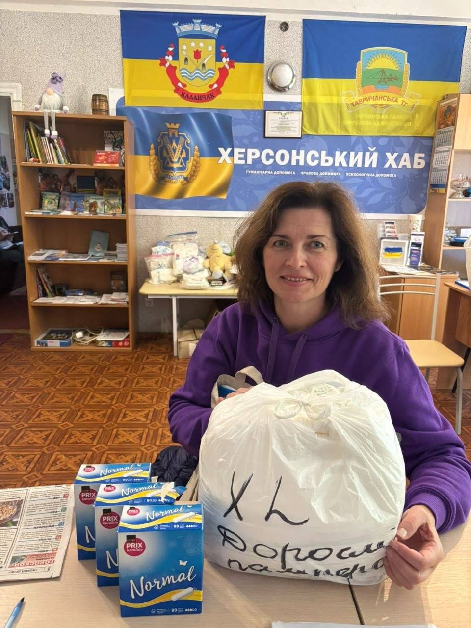 Усміхнена жінка-переселенка в яскравому фіолетовому худі тримає в руках великий білий пакунок із гуманітарною допомогою в приміщенні координаційного центру «Вільні разом» у Києві. На мішку зроблено чорний напис «XL Дорослі памперси», а на передньому плані на столі з візерунчастою поверхнею розкладено кілька пачок синіх гігієнічних засобів Prix «Normal» та свіжий примірник друкованої газети. Видача допомоги для внутрішньо переміщених осіб із Херсонщини проходить у робочому кабінеті, де на задньому плані встановлено великий синій банер «ХЕРСОНСЬКИЙ ХАБ» із підписами про надання гуманітарної, правової та психологічної допомоги, над яким на стіні закріплено жовто-блакитні прапори Каланчацької та Тавричанської громад із їхніми офіційними гербами. Ліворуч у кімнаті височіє дерев'яна шафа-стелаж із книгами, журналами, іграшковим гномом на полиці, а на тумбі по центру складено додаткові пакунки, дитячі речі та засоби гігієни для нужденних.