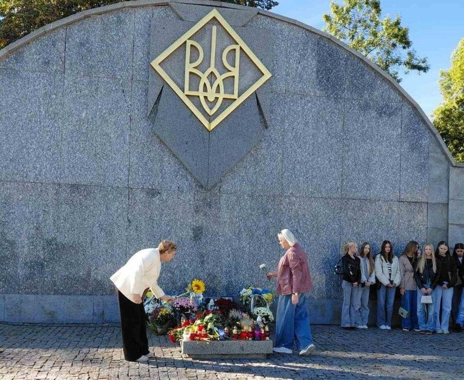 Дві жінки похилого віку з числа переселенок із Херсонщини схиляються перед гранітним меморіалом і покладають квіти під час заходів до Дня Захисників і Захисниць України, організованих координаційним центром «Вільні разом» у Львові на Марсовому полі. На передньому плані ліворуч учасниця зустрічі у світлому жакеті та чорних штанях акуратно поправляє букет біля підніжжя монумента, поруч із нею інша жінка у рожевій куртці та широких синіх джинсах тримає білу квітку, готуючись покласти її поруч із лампадками, живими соняшниками та трояндами. Урочисте вшанування пам'яті полеглих воїнів проходить біля високої дугоподібної стіни з сірого граніту, у верхній частині якої встановлено великий золотий тризуб у ромбі, водночас праворуч уздовж меморіального знака на вимощеній бруківкою площі вишикувалася група молодих дівчат, які уважно спостерігають за процесом під яскравим сонцем на тлі зелених дерев.