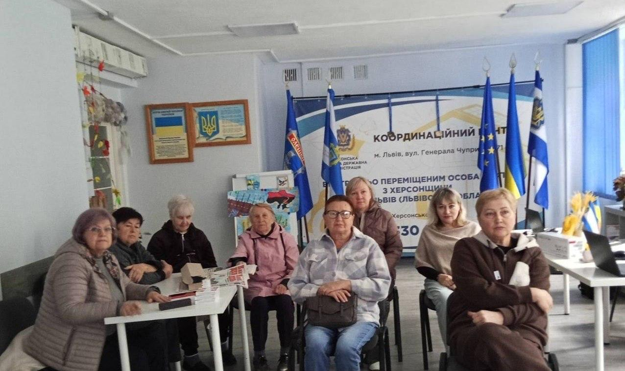 Група переселенок із Херсонщини позує для спільного пам'ятного фото у світлому робочому кабінеті координаційного центру «Вільні разом» у Львові. Жінки розсаджені півколом, де ліворуч частина учасниць перебуває за білим столом із друкованими інформаційними матеріалами та газетами, а інші присутні сидять по центру та праворуч у кімнаті, уважно дивлячись у кадр. Зустріч проходить на тлі великого офіційного банера хабу, з лівого боку від якого на флагштоках піднято синій прапор із написом «Олешки» та Державний прапор України, а з правого боку встановлено прапор Європейського Союзу, Державний прапор України та прапор Херсонської області. На дальньому плані ліворуч на стіні закріплено інформаційні стенди з державною символікою України, під ними виставлено дитячі малюнки, а праворуч у залі помітно край білого робочого столу з відкритим ноутбуком та вікно з блакитними вертикальними жалюзі.