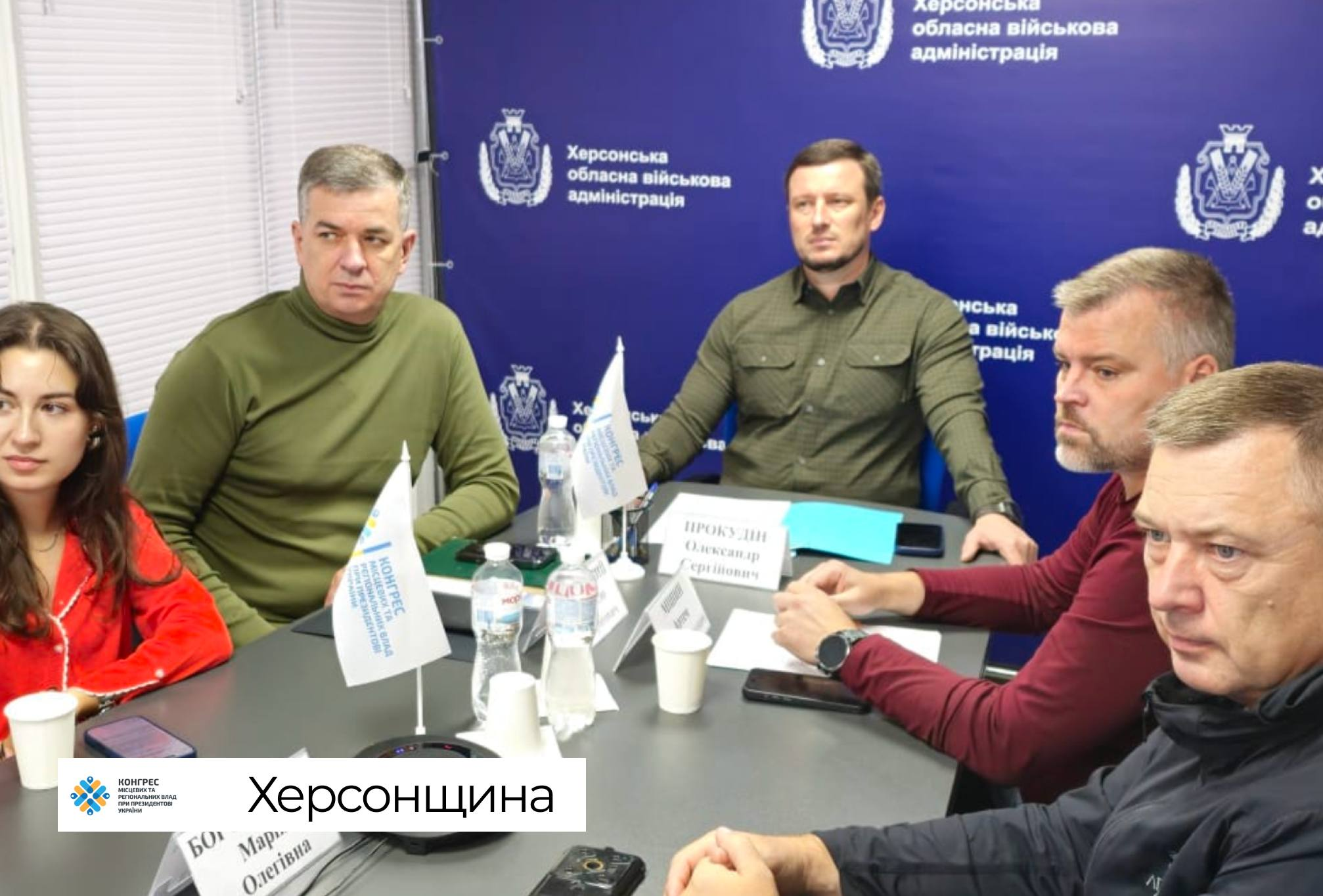 Керівництво Херсонської ОВА та МВА разом із колегами сидить за робочими столами під час участі у фінальній стратегічній сесії Офісу Конгресу місцевих та регіональних влад при Президентові України, яка присвячена публічним інвестиціям та наповненню місцевих бюджетів громад. На передньому плані ліворуч сидить начальниця управління міжнародного співробітництва та залучення інвестицій ХОДА (Офіс інвестицій та розвитку експорту) Марія Богодухова в червоній кофті перед табличкою зі своїм іменем, праворуч від неї розмістився перший заступник начальника Херсонської МВА Михайло Линецький в оливковому світшоті, а далі по центру за столом перебуває начальник Херсонської обласної військової адміністрації Олександр Прокудін у сорочці захисного кольору перед іменним підписом та мобільним телефоном. Безпосередньо праворуч від Олександра Прокудіна сидить директор Департаменту розвитку економіки Херсонської обласної державної адміністрації Артем Мішин у темно-бордовому світшоті, біля якого в ряд розмістилися інші учасники робочої зустрічі. На білих столах розставлено пляшки мінеральної води, паперові стаканчики, блокноти для записів та встановлено маленькі настільні прапорці з логотипом Конгресу місцевих та регіональних влад, а уся офіційна нарада проходить на тлі великого синього брендованого банера із зображенням офіційного герба Херсонської області та білими написами «Херсонська обласна військова адміністрація».