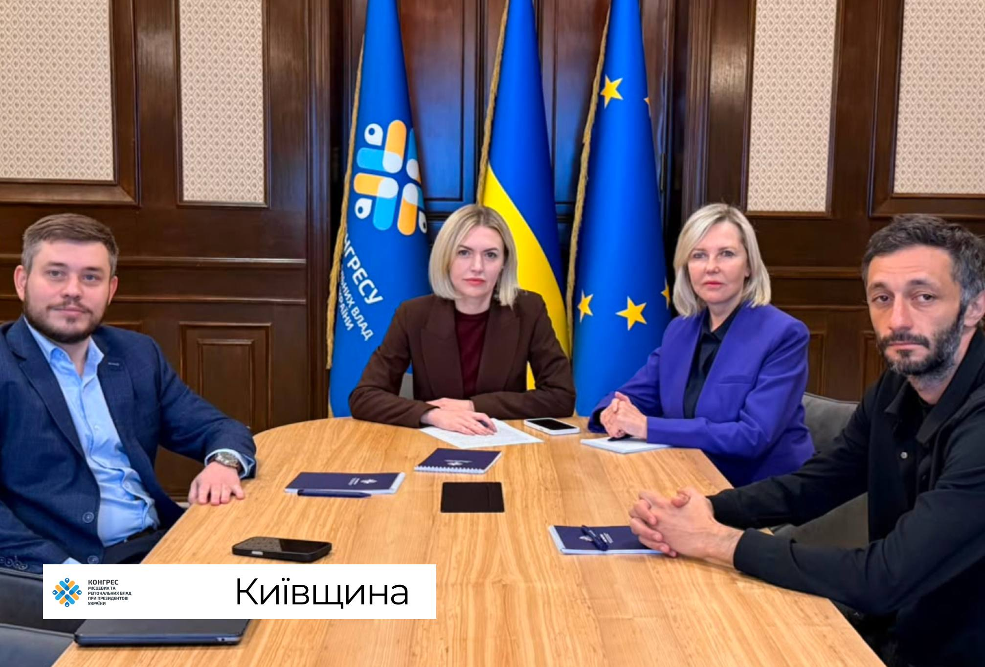 Керівництво Київської обласної державної адміністрації сидить за великим дерев'яним столом під час онлайн-участі у фінальній стратегічній сесії Офісу Конгресу місцевих та регіональних влад при Президентові України щодо розвитку та наповнення бюджетів громад. На передньому плані ліворуч сидить чоловік у синьому піджаку та світлій сорочці, по центру за столом розмістилися дві жінки — одна в коричневому блейзері поверх темного топа, а поруч із нею її колега в яскраво-синьому піджаку, водночас праворуч сидить чоловік у чорній сорочці зі складеними в замок руками. На столі перед присутніми розкладено сині блокноти з ручками та мобільні телефони, а офіційна робоча зустріч проходить у кабінеті з класичними темними дерев'яними стіновими панелями на тлі трьох піднятих на флагштоках великих стягів: синього прапора Конгресу місцевих та регіональних влад, Державного прапора України та прапора Європейського Союзу.