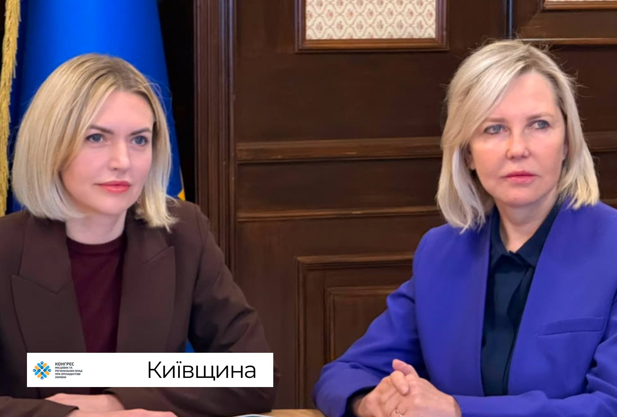 Дві посадовиці Київської обласної державної адміністрації зосереджено працюють за великим дерев'яним столом під час онлайн-участі у фінальній стратегічній сесії Офісу Конгресу місцевих та регіональних влад при Президентові України, присвяченій розвитку та наповненню бюджетів громад. Ліворуч у кадрі в коричневому блейзері поверх темного топа сидить перша представниця керівництва, а праворуч від неї розмістилася її колега в яскраво-синьому піджаку, тримаючи пальці рук складеними в замок. Офіційне засідання, спрямоване на аналіз економіки, виявлення фінансових резервів та посилення спроможності територіальних громад, проходить у робочому кабінеті з класичними темними дерев'яними стіновими панелями, де ліворуч на задньому плані частково видно піднятий синій прапор Конгресу місцевих та регіональних влад.