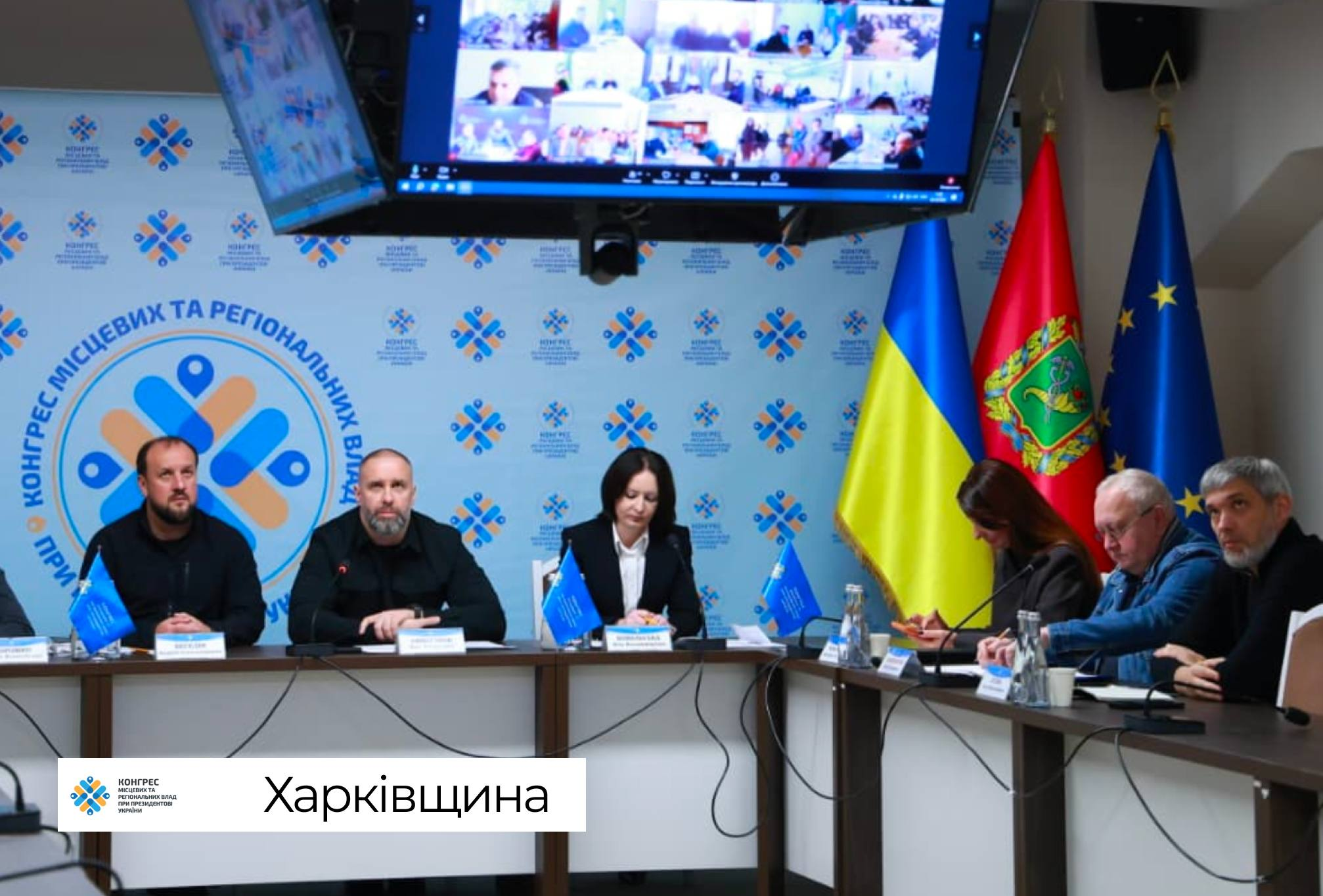 Представники керівництва Харківської обласної державної адміністрації сидять за великим робочим столом пресцентру під час проведення фінальної стратегічної сесії Офісу Конгресу місцевих та регіональних влад при Президентові України, яка присвячена публічним інвестиціям та наповненню місцевих бюджетів громад. За центральною частиною столу ліворуч сидить посадовець у чорному худі, поруч із ним по центру перебуває його колега у чорному світшоті, а праворуч розмістилася учасниця наради у чорному піджаку та білій блузці. З правого боку столу в ряд сидить інша група фахівців, які роблять записи у блокнотах, водночас на столах перед присутніми розставлено пляшки мінеральної води, паперові стаканчики та встановлено маленькі сині настільні прапорці з емблемою заходу. Уся офіційна зустріч проходить на тлі великого світло-блакитного брендованого банера з круглим логотипом і написом «КОНГРЕС МІСЦЕВИХ ТА РЕГІОНАЛЬНИХ ВЛАД ПРИ ПРЕЗИДЕНТОВІ УКРАЇНИ», над яким під стелею підвішено великий телевізійний екран із сіткою онлайн-трансляції інших регіонів, а у правому кутку залу на флагштоках піднято Державний прапор України, червоний прапор Харківської області з гербом та прапор Європейського Союзу.