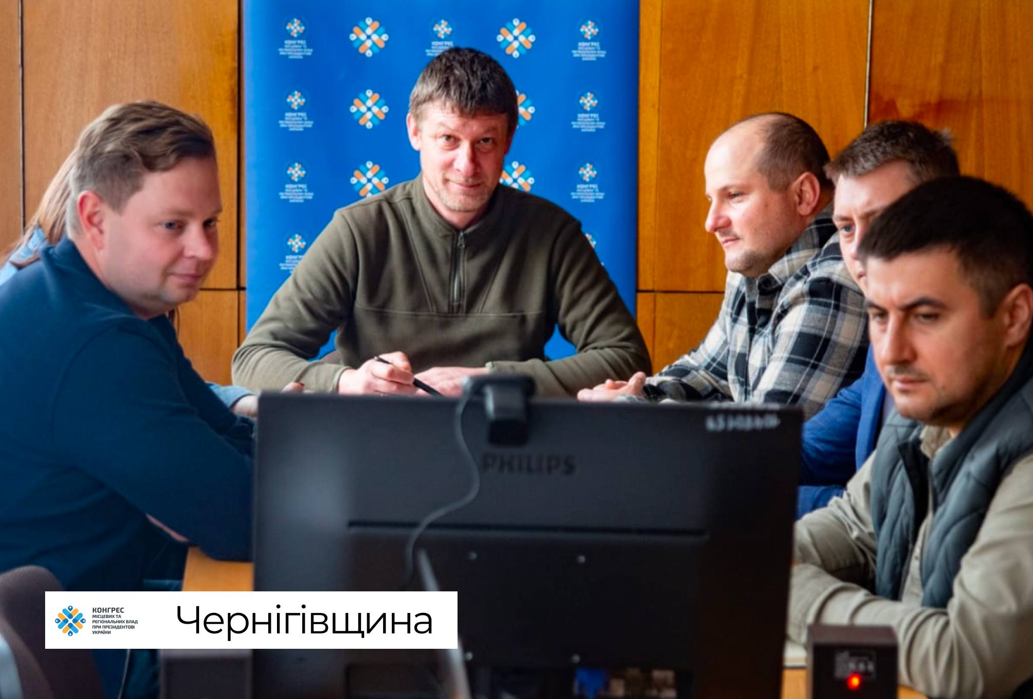 Представники керівництва Чернігівської обласної державної адміністрації сидять за робочим столом перед екраном великого чорного монітора Philips під час онлайн-участі у фінальній стратегічній сесії Офісу Конгресу місцевих та регіональних влад при Президентові України. З лівого краю кадру сидить учасник наради у темно-синьому светрі, повернувши голову в профіль, поруч із ним по центру перебуває колега в оливковому худі на блискавці, який тримає в руках ручку, а праворуч у ряд розмістилися інші посадовці, зокрема чоловік у сорочці в чорно-білу клітинку, фахівець у синьому піджаку та учасник у зеленій куртці-жилетці на передньому плані. Робоча зустріч, присвячена аналізу економічних показників, публічним інвестиціям та наповненню місцевих бюджетів громад області, проходить у кабінеті з дерев'яними стіновими панелями на тлі синього вертикального інформаційного банера Офісу Конгресу з його фірмовими біло-помаранчево-блакитними емблемами.