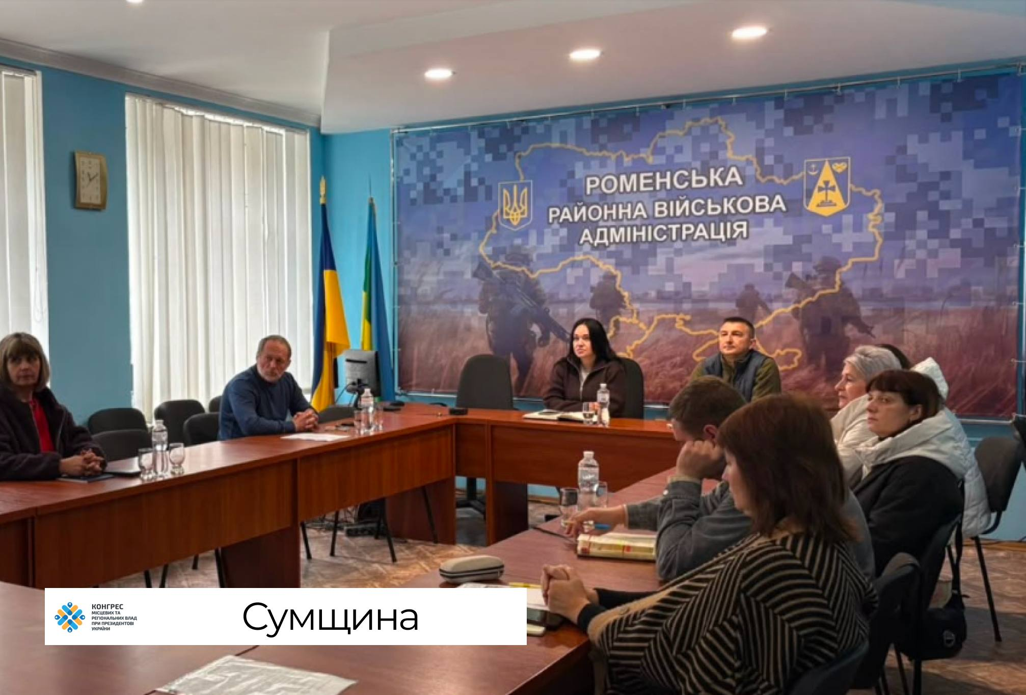 Представники Сумщини беруть участь у фінальній стратегічній сесії Офісу Конгресу місцевих та регіональних влад при Президентові України, сидітимуть за великим дерев'яним столом у формі літери П. Робоча зустріч для аналізу економіки та наповнення місцевих бюджетів громад проходить у залі засідань, де на передньому плані праворуч учасники наради роблять нотатки в блокнотах і працюють із документами. На дальньому плані у центрі столу перебувають керівники, а ліворуч за окремою секцією столу сидять інші представники Сумської області біля вікна з вертикальними жалюзі, де в кутку кімнати встановлено на флагштоках Державний прапор України та зелений прапор. На столах розставлено пляшки з мінеральною водою та склянки, а вся офіційна зустріч проходить на тлі великого тематичного банера Роменської районної військової адміністрації із зображенням контурної карти Сумщини, офіційного тризуба, герба району та силуетів українських захисників на полі.