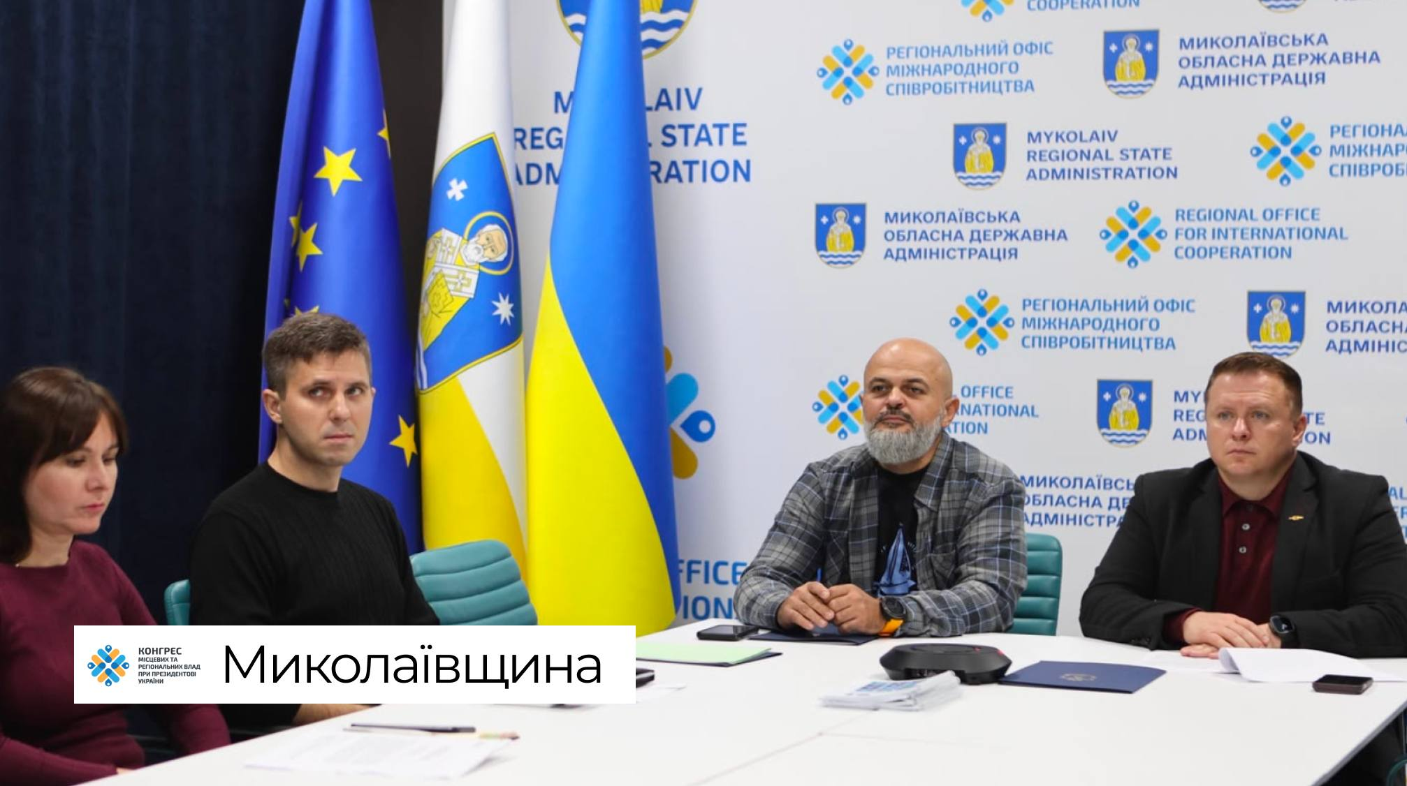 Представники Миколаївщини беруть участь у фінальній стратегічній сесії Офісу Конгресу місцевих та регіональних влад при Президентові України, перебуваючи за світлим робочим столом у залі засідань. З лівого краю кадру сидить учасниця наради в бордовій кофті, поруч із якою по центру розмістився лисий чоловік із бородою в сорочці в сіро-чорну клітинку, а праворуч сидить його колега в темному піджаку та бордовому світшоті під ним, тримаючи руки перед собою. На передньому плані перед присутніми розкладено папки з паперами, блокноти для записів та мобільні телефони, водночас офіційна робоча зустріч, спрямована на аналіз економіки, публічних інвестицій та наповнення місцевих бюджетів громад, проходить на тлі великого світлого брендованого банера Регіонального офісу міжнародного співробітництва Миколаївської обласної державної адміністрації з його численними фірмовими синьо-жовтими емблемами та написами українською й англійською мовами, ліворуч від якого на високих флагштоках піднято прапор Європейського Союзу, синьо-білий прапор Миколаївської області з гербом святого Миколая та Державний прапор України.