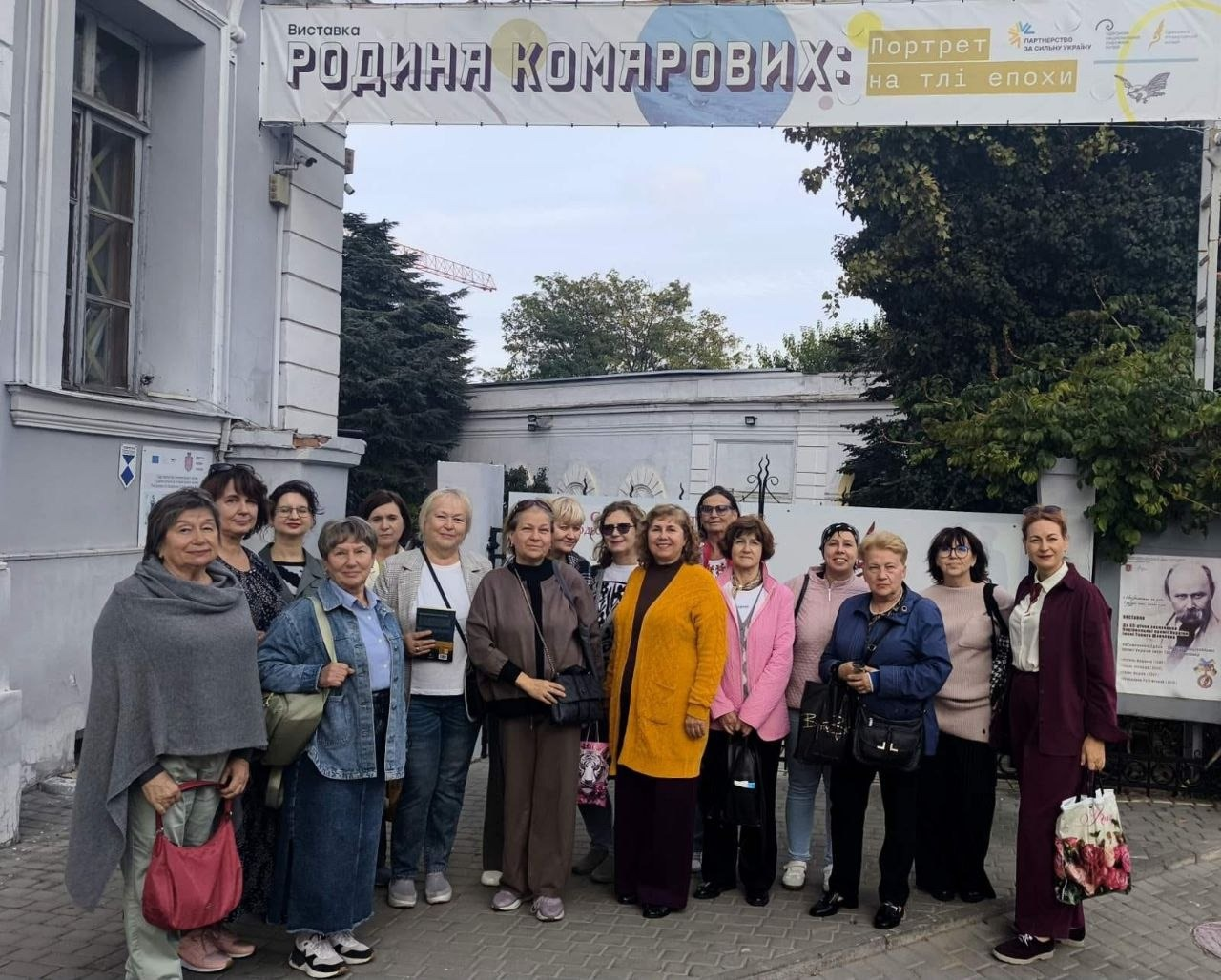 Група переселенок із Херсонщини позує для спільного пам'ятного фото на вимощеному бруківкою подвір'ї перед входом до Одеського літературного музею під час культурно-просвітницького заходу, організованого публічною бібліотекою Верхньорогачицької селищної ради. Жінки різного віку у повсякденному осінньому одязі, куртках, пальтах та светрах вишикувалися разом під великим білим банером із написами «Виставка РОДИНА КОМАРОВИХ: Портрет на тлі епохи», який закріплений над арковим проходом будівлі. Серед екскурсанток на передньому плані ліворуч стоїть жінка у сірому довгому кардигані з червоною сумкою через плече, поруч із нею — учасниця у джинсовій куртці з бежевим рюкзаком, по центру виділяється жінка в яскравій подовженій жовтій кофті, а з правого краю помітна відвідувачка у класичному темно-бордовому костюмі. На задньому плані за спинами присутніх видно білі стіни музейного комплексу, металеві декоративні ворота, густі зелені дерева, а праворуч на паркані закріплена інформаційна афіша заходу з портретним малюнком.