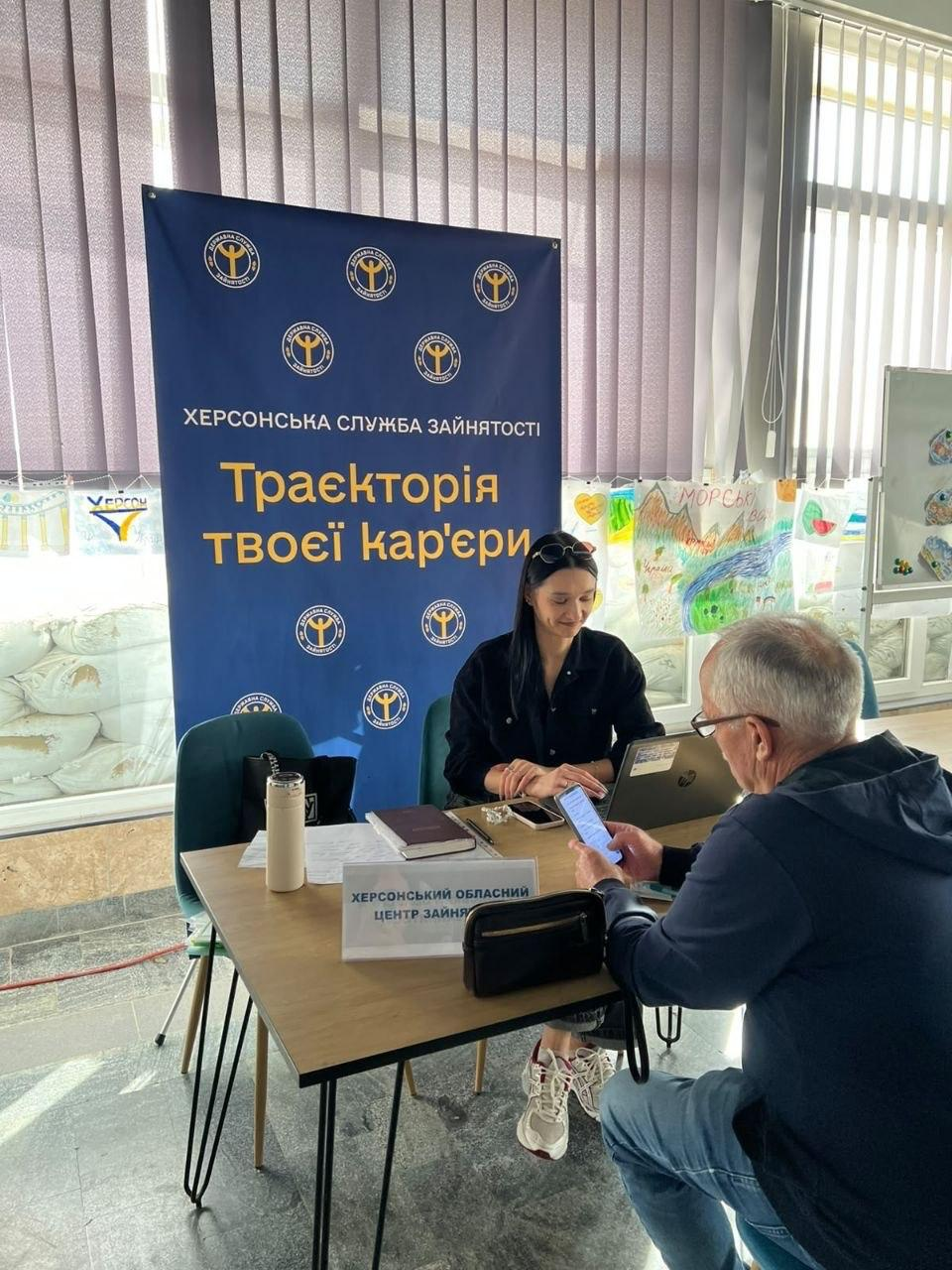 Співробітниця Херсонської служби зайнятості у чорній сорочці сидить за робочим столом та надає індивідуальну консультацію літньому переселенцю в синьому худі, який тримає в руці мобільний телефон, під час заходу з комплексної підтримки ВПО у миколаївському координаційному центрі «Вільні разом». Консультантка уважно дивиться на екран смартфона відвідувача, при цьому на столі перед нею відкритий сріблястий ноутбук, лежить щоденник, а на передньому плані встановлено настільну табличку «ХЕРСОНСЬКИЙ ОБЛАСНИЙ ЦЕНТР ЗАЙНЯТОСТІ». Робоча зустріч для кар'єрного консультування та допомоги у працевлаштуванні херсонців проходить у світлому приміщенні на тлі великого синього банера з офіційними золотими емблемами та великими написами «ХЕРСОНСЬКА СЛУЖБА ЗАЙНЯТОСТІ» та «Траєкторія твоєї кар'єри», а за ним на стіні під жалюзі вивішено виставку дитячих малюнків.