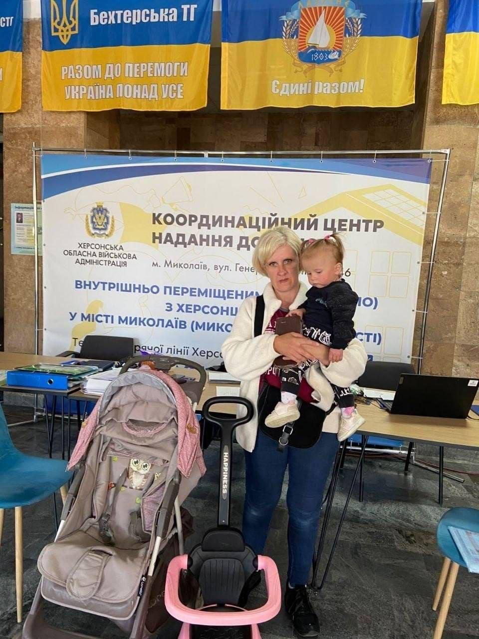 Жінка-переселенка з Херсонщини у світлій куртці та синіх джинсах тримає на руках маленьку донечку, отримуючи дитячий візочок та допоміжні засоби для дитини від благодійного фонду «Медицина Херсонщини» у миколаївському координаційному центрі «Вільні разом». Мати з дитиною стоїть на повний зріст по центру кадру перед робочими столами, де ліворуч від неї розміщено сучасний прогулянковий дитячий візочок сіро-рожевого кольору, а попереду виставлено дитяче сидіння-самокат чорно-рожевого кольору з написом «HAPPINESS» на ручці. Гуманітарна допомога та підтримка сімей ВПО проходить у приміщенні на тлі великого офіційного банера «КООРДИНАЦІЙНИЙ ЦЕНТР НАДАННЯ ДОПОМОГИ ВНУТРІШНЬО ПЕРЕМІЩЕНИМ ОСОБАМ З ХЕРСОНЩИНИ У МІСТІ МИКОЛАЇВ», над яким на стіні закріплено жовто-блакитні прапори Бехтерської територіальної громади з патріотичними гаслами «РАЗОМ ДО ПЕРЕМОГИ. УКРАЇНА ПОНАД УСЕ» та «Єдині разом!».