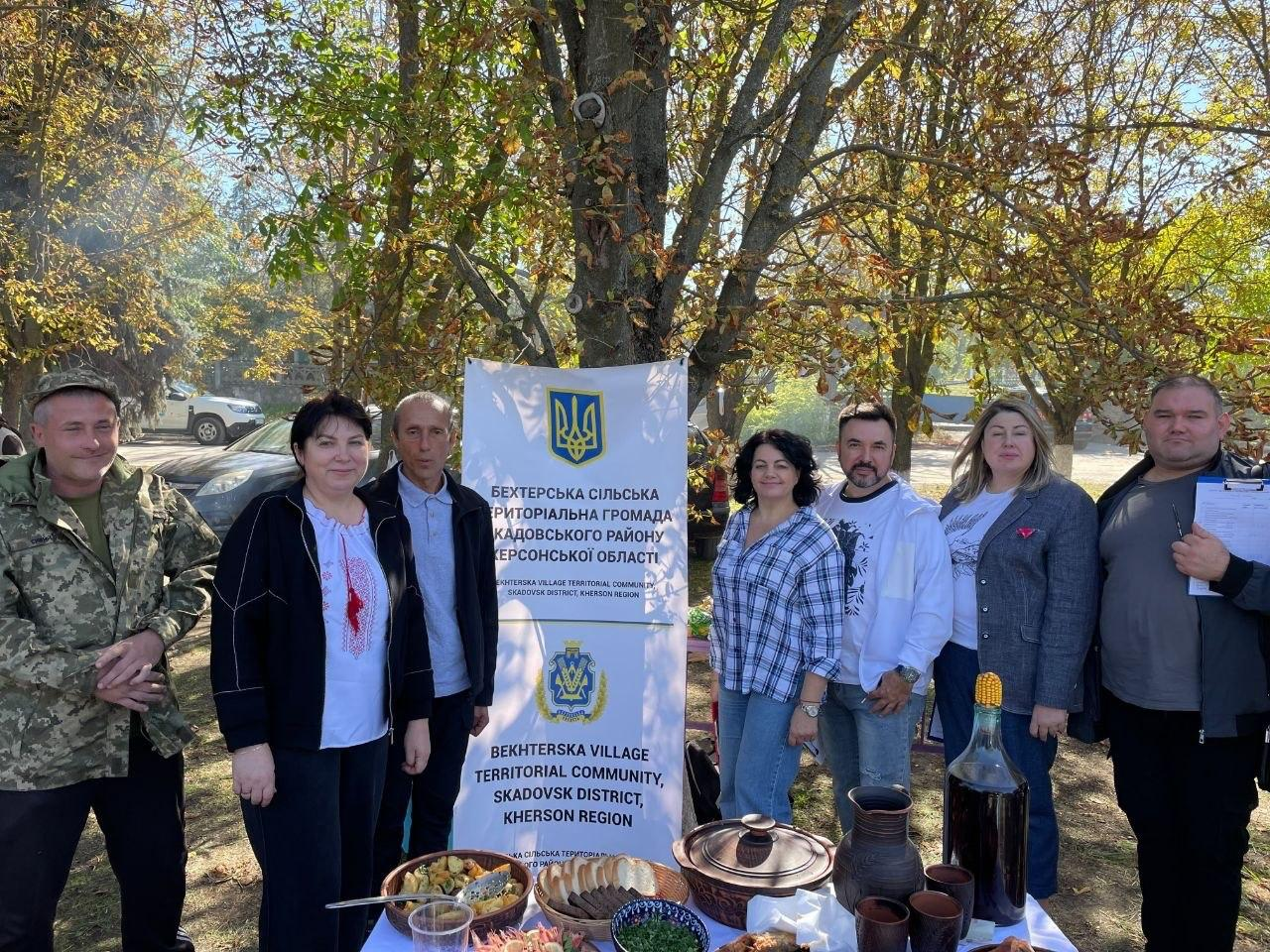 Група представників Бехтерської громади та координаційного центру «Вільні разом» позує для спільного пам'ятного фото навколо святкового столу зі стравами традиційної української кухні під час участі у благодійному фестивалі-ярмарку «Смакуємо в рідному селі» на підтримку ЗСУ. Учасники заходу вишикувалися в один ряд на тлі осінніх дерев та припаркованих автомобілів, розділившись по обидва боки від високого вертикального білого банера із зображенням Державного Герба України та двомовним написом «БЕХТЕРСЬКА СІЛЬСЬКА ТЕРИТОРІАЛЬНА ГРОМАДА СКАДОВСЬКОГО РАЙОНУ ХЕРСОНСЬКОЇ ОБЛАСТІ». З лівого боку від інформаційного стенда стоять військовослужбовець у камуфляжній формі та кепці, жінка у чорному кардигані з вишитим червоним орнаментом на білій блузці та чоловік у класичному синьому костюмі зі світлою сорочкою. Праворуч від банера розмістилися жінка у картатій синьо-білій сорочці, чоловік у білому світшоті з патріотичним малюнком, жінка у сірому піджаку з червоною стрічкою-оберегом на грудях та чоловік у сірій кофті, який тримає в руках планшет із паперами для записів. На передньому плані перед присутніми облаштовано стіл із білою скатертиною, де виставлено великі глиняні горщики, плетену миску з нарізаним домашнім хлібом, тарілки з традиційними частуваннями, глиняні куманці та велику прозору пляшку, яка запечатана качаном кукурудзи.