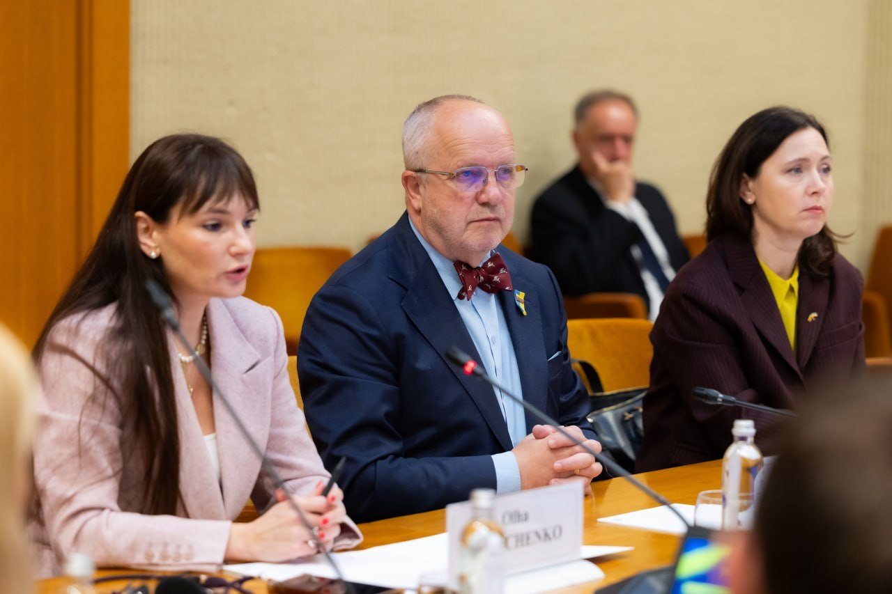 Віцеспікер Сейму Литовської Республіки Юозас Олекас разом із колегами сидить за робочим столом президії під час проведення міжнародної конференції «Укриття: досвід України та рішення Литви». З лівого краю кадру сидить представниця литовської сторони в рожевому жакеті, яка виступає перед присутніми у залі з мікрофоном, по центру розмістився Юозас Олекас у синьому піджаку, світлій сорочці та фірмовому темно-червоному метелику з окулярами на обличчі, а праворуч від нього перебуває заступниця міністра внутрішніх справ Литви Ана Бурковскєнє у темно-бордовому костюмі та жовтій блузці. На дерев'яному столі перед учасниками засідання встановлено настільні мікрофони, розкладено паперові документи, блокноти для записів та виставлено іменні таблички, а на задньому плані кадру на тлі світлої стіни залу засідань помітно силует іншого учасника наради у чорному костюмі та білій сорочці.