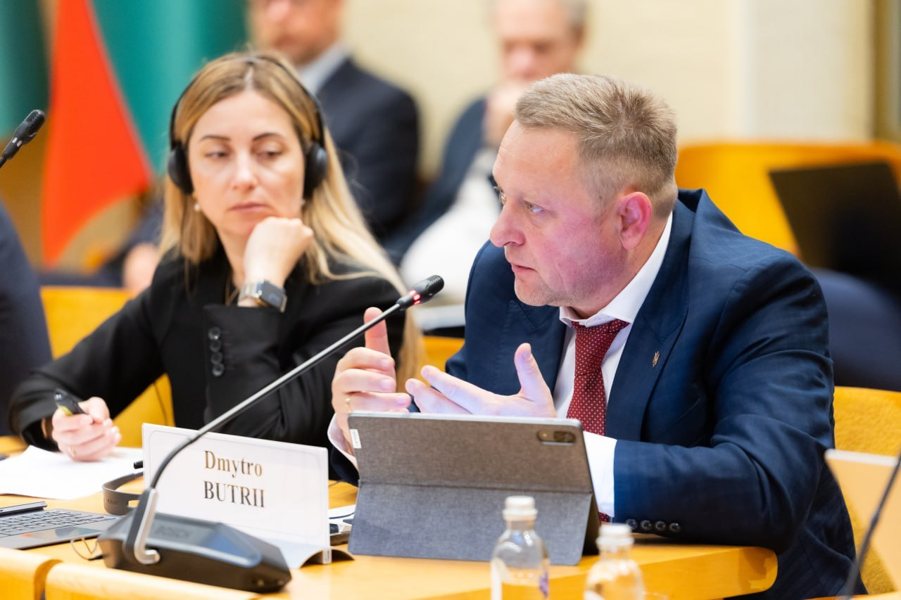 Перший заступник начальника Херсонської обласної військової адміністрації Дмитро Бутрій виступає з промовою перед учасниками міжнародної конференції «Укриття: досвід України та рішення Литви» у залі засідань литовського Сейму. Посадовець у темно-синьому костюмі, білій сорочці та червоній краватці сидить праворуч за дерев'яним столом, активно жестикулюючи руками та детально розповідаючи про практичний досвід створення мережі безпечних просторів і розширення фонду захисних споруд на деокупованій Херсонщині. Перед ним на столі встановлено сірий планшет у чохлі-підставці, пляшку мінеральної води, настільний мікрофон Bosch та велику іменну білу табличку з написом «Dmytro BUTRII». Ліворуч від нього в навушниках синхронного перекладу сидить заступниця директора Агенції регіонального розвитку Херсонщини Інна Бутрій у чорному жакеті, яка уважно слухає виступ та тримає в руці ручку перед відкритим ноутбуком, а на дальньому плані за їхніми спинами помітно силуети інших експертів та піднятий Державний прапор Литви.