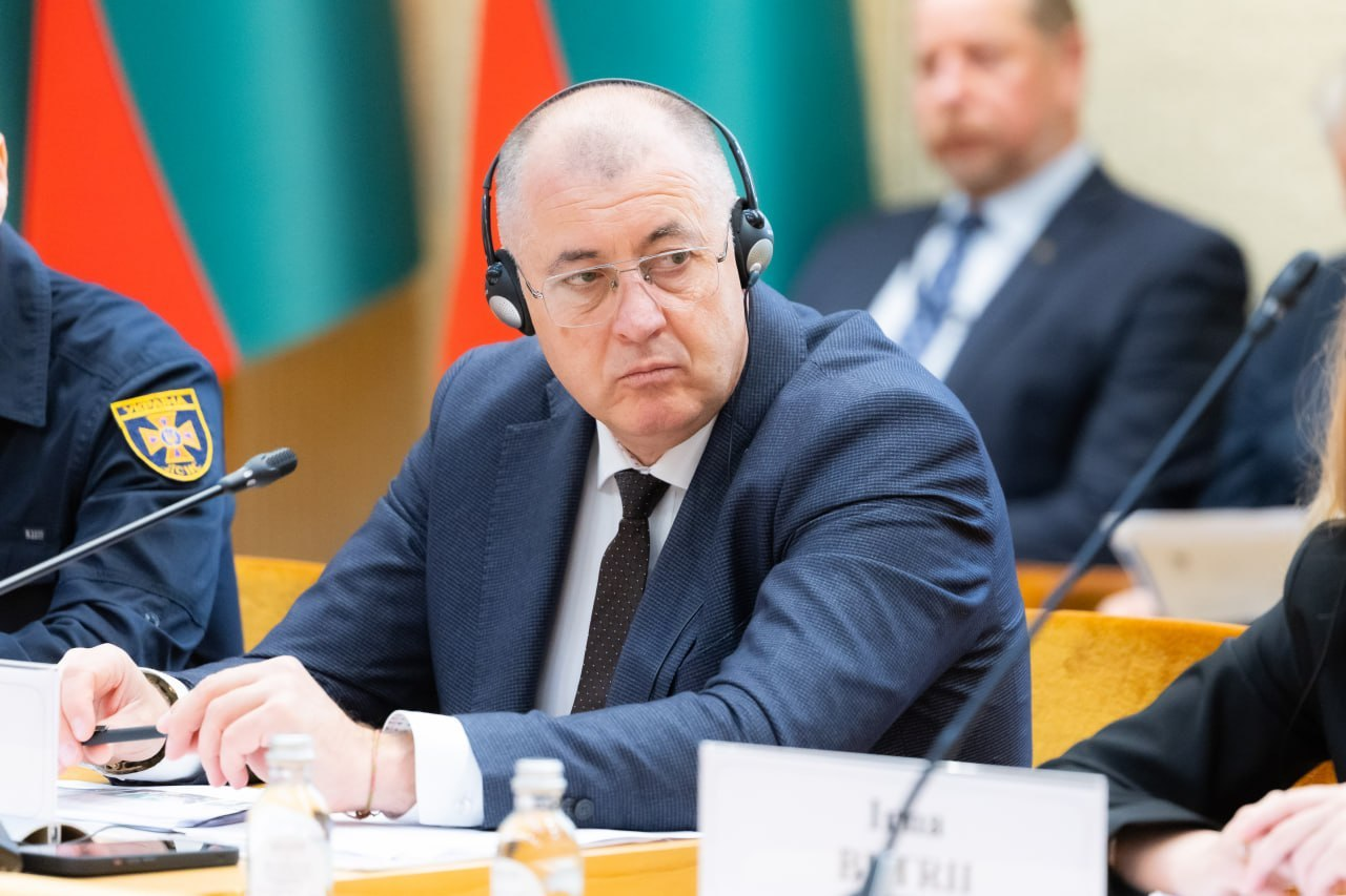 Начальник Дар’ївської сільської військової адміністрації Анатолій Дідур в окулярах та навушниках синхронного перекладу уважно слухає виступи колег під час міжнародної конференції «Укриття: досвід України та рішення Литви» у залі засідань литовського Сейму. Посадовець у класичному темно-синьому костюмі, білій сорочце та темній краватці сидить по центру за дерев'яним робочим столом, де перед ним розкладено папери, блокноти для записів та встановлено настільний мікрофон. Праворуч від нього розмістилася заступниця директора Агенції регіонального розвитку Херсонщини Інна Бутрій, перед якою на столі встановлено іменну білу табличку з написом «Inna BUTRII», а ліворуч у кадрі сидить представник Державної служби України з надзвичайних ситуацій у чорній форменій сорочці з офіційним шевроном ДСНС України на рукаві. На дальньому плані за спинами присутніх на тлі світлої стіни залу засідань помітно державного службовця у діловому костюмі та вертикальні смуги двох піднятих Державних прапорів Литви.