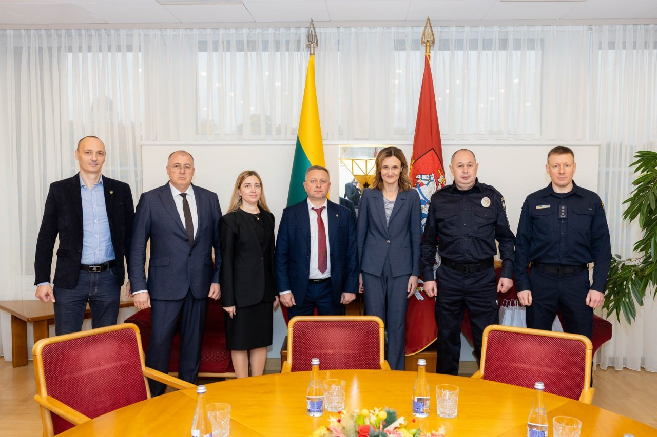 Представники української делегації на чолі з першим заступником начальника Херсонської ОВА Дмитром Бутрієм та литовські посадовці стоять у спільній шерензі під час міжнародної конференції «Укриття: досвід України та рішення Литви» в Сеймі Литовської Республіки. Учасники зустрічі вишикувалися в один ряд на тлі великих вікон із білими вертикальними жалюзі та піднятих на високих дерев'яних флагштоках Державного прапора Литовської Республіки й червоного прапора з історичним литовським гербом Погоня. Якщо дивитися на знімок зліва направо, у залі стоять: чоловік у синій сорочці та чорному піджаку, Віцеспікер Сейму Литовської Республіки Юозас Олекас у класичному темному костюмі та краватці, заступниця директора Агенції регіонального розвитку Херсонщини Інна Бутрій у чорному костюмі, перший заступник начальника Херсонської ОВА Дмитро Бутрій у синьому костюмі та червоній краватці, віцеспікерка Сейму Вікторія Чміліте-Нільсен у темно-синьому жакеті, а також двома останніми з правого краю стоять українські правоохоронці у чорній форменій сорочці та синьому однострої поліції. На передньому плані перед присутніми видно край великого круглого лакованого столу з квітковою композицією в центрі та розставленими скляними пляшками мінеральної води й прозорими стаканчиками, а навколо столу розставлені дерев'яні крісла з м'якою бордовою оббивкою.