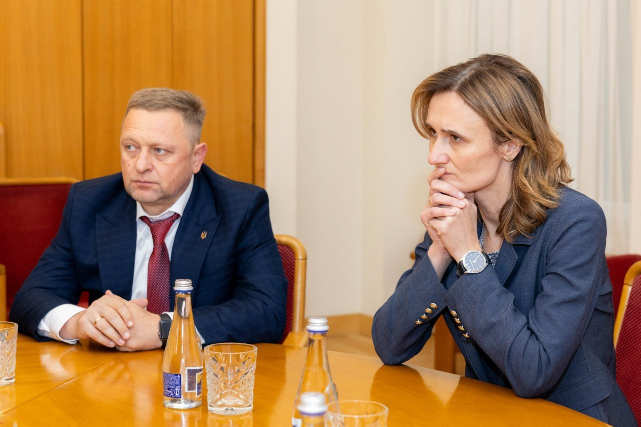Перший заступник начальника Херсонської обласної військової адміністрації Дмитро Бутрій та віцеспікерка Сейму Литовської Республіки Вікторія Чміліте-Нільсен сидять поруч за дерев'яним столом під час зустрічі у межах міжнародної конференції «Укриття: досвід України та рішення Литви». Ліворуч у кадрі Дмитро Бутрій у класичному темно-синьому костюмі, білій сорочці та червоній краватці тримає руки на столі із замкненими пальцями й уважно слухає виступи колег під час обговорення безпекових потреб громад. Праворуч від нього розмістилася Вікторія Чміліте-Нільсен у темно-синьому жакеті з наручним годинником, яка зосереджено дивиться перед собою, спершись підборіддям на складені руки. Перед посадовцями на полірованій поверхні столу розставлено прозорі скляні пляшки з мінеральною водою та кришталеві стаканчики, а сама робоча зустріч щодо розширення фонду захисних споруд прифронтових регіонів України проходить у світлому кабінеті Сейму на тлі нейтральної бежевої стіни та дерев'яних міжкімнатних дверей.