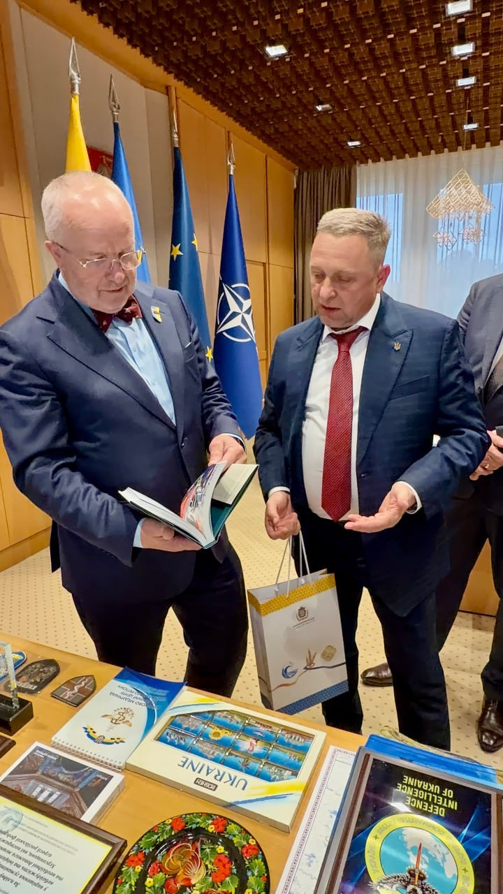 Перший заступник начальника Херсонської обласної військової адміністрації Дмитро Бутрій спілкується з Віцеспікером Сейму Литовської Республіки Юозасом Олекасом біля столу з пам'ятними подарунками під час міжнародної конференції «Укриття: досвід України та рішення Литви». Ліворуч у кадрі Юозас Олекас у синьому костюмі та бордовому метелику уважно розглядає та гортає розгорнуту книгу, тоді як праворуч Дмитро Бутрій у темно-синьому костюмі, білій сорочці та червоній краватці тримає в руці подарунковий паперовий пакет із державною символікою та веде розмову. На столі перед посадовцями виставлено пам'ятні сувеніри, серед яких велика подяка у рамці, декоративна тарілка з народним розписом, книга з логотипом Головного управління розвідки «DEFENCE INTELLIGENCE OF UKRAINE» та набір поштових марок «UKRAINE». Офіційна зустріч щодо розширення фонду захисних споруд і допомоги прифронтовим громадам проходить у світлому кабінеті Сейму на тлі ряду піднятих великих стягів: Державного прапора Литовської Республіки, Державного прапора України, прапора Європейського Союзу та прапора НАТО.