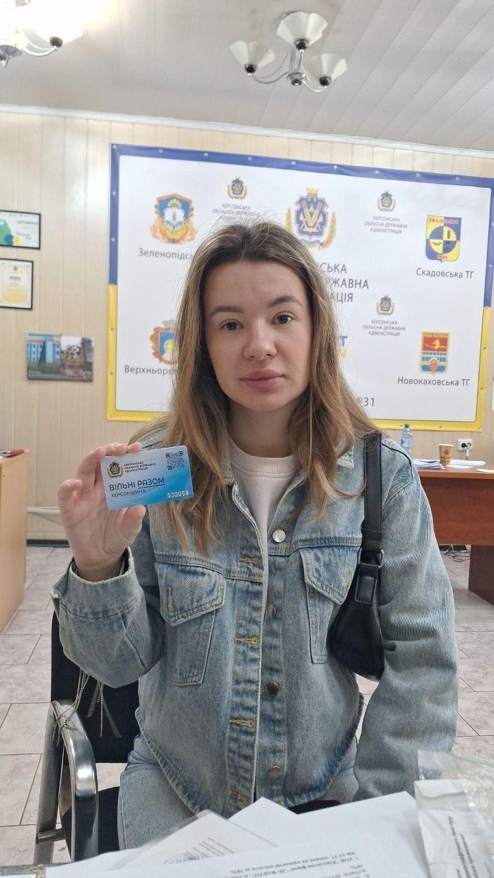 Молода жінка-переселенка з Херсонщини у блакитній джинсовій куртці та світлій кофті сидить у приміщенні хабу й демонструє перед собою пластикову «Картку Херсонщини» під час заходу з підтримки ВПО від координаційного центру «Вільні разом» та БФ «Сапорт Херсон» в Одесі. Вона тримає у правій руці невелику синьо-блакитну картку з офіційним гербом, QR-кодом та великим написом «ВІЛЬНІ РАЗОМ ХЕРСОНЩИНА», а на її лівому плечі висить тонка чорна сумочка-багет. Видача карток для внутрішньо переміщених осіб проходить у робочому кабінеті на тлі великого світлого банера з емблемами Херсонської ОВА та гербами громад, серед яких чітко видно символіку Зеленопідської, Верхньорогачицької, Скадовської та Новокаховської територіальних громад, при цьому ліворуч на стіні висять грамоти в рамках, а праворуч облаштовано дерев'яні столи з канцелярією.