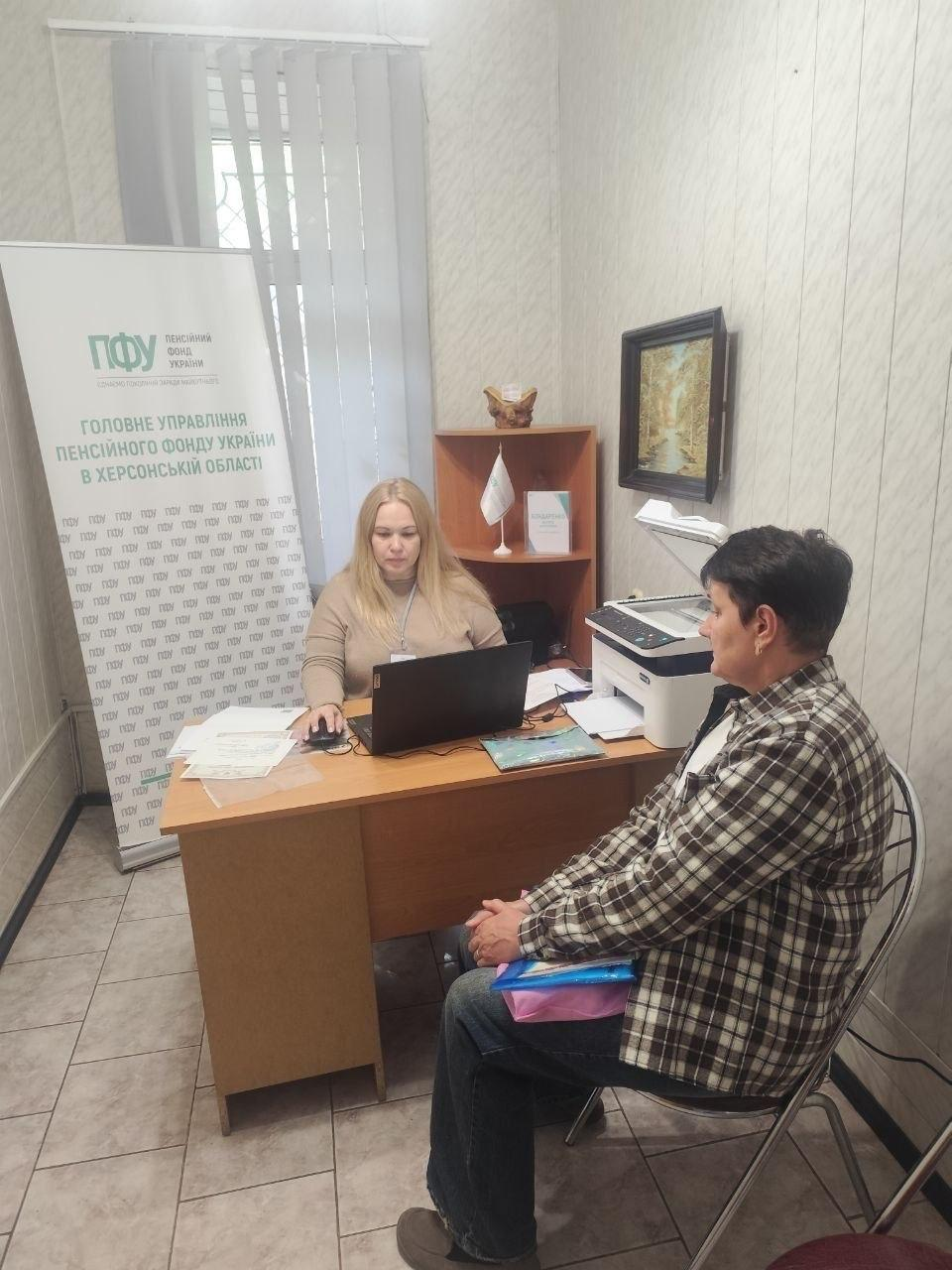 Фахівчиня Головного управління Пенсійного фонду України в Херсонській області надає індивідуальну консультацію з пенсійних питань жінці-переселенці у приміщенні одеського координаційного центру «Вільні разом». Співробітниця ПФУ в бежевій кофті сидить за дерев'яним кутовим столом і працює за відкритим чорним ноутбуком, водночас перед нею у профіль до кадру сидить відвідувачка у картатій чорно-білій сорочці та джинсах, яка тримає на колінах синю папку з документами. На столі біля фахівчині розкладено папери, а праворуч на тумбі встановлено білий багатофункціональний принтер-сканер, над яким на стіні висить картина у темній рамці. Індивідуальний прийом громадян для всебічної допомоги ВПО, організований спільно з БФ «Сапорт Херсон», проходить у робочому кабінеті на тлі вертикальних білих жалюзі та великого білого інформаційного банера з фірмовим зеленим логотипом «ПФУ» та великим написом «ГОЛОВНЕ УПРАВЛІННЯ ПЕНСІЙНОГО ФОНДУ УКРАЇНИ В ХЕРСОНСЬКІЙ ОБЛАСТІ».