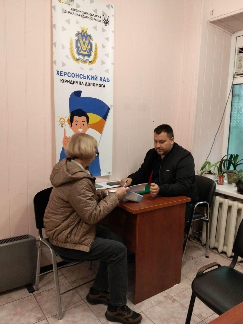 Юрист у чорній куртці сидить за дерев'яним столом та надає індивідуальну правову консультацію жінці-переселенці у приміщенні одеського хабу, організовану в межах комплексної підтримки ВПО від координаційного центру «Вільні разом» та БФ «Сапорт Херсон». Консультант тримає в руці ручку й вивчає надані документи, водночас перед ним у профіль до об'єктива сидітиме відвідувачка в окулярах та бежевій куртці, яка уважно слухає роз'яснення фахівця. Робоча зустріч для правового захисту громадян проходить у кабінеті на тлі великого білого вертикального банера з офіційним гербом, написом «ХЕРСОНСЬКИЙ ХАБ. ЮРИДИЧНА ДОПОМОГА» та патріотичним малюнком хлопчика із синьо-жовтим прапором України.