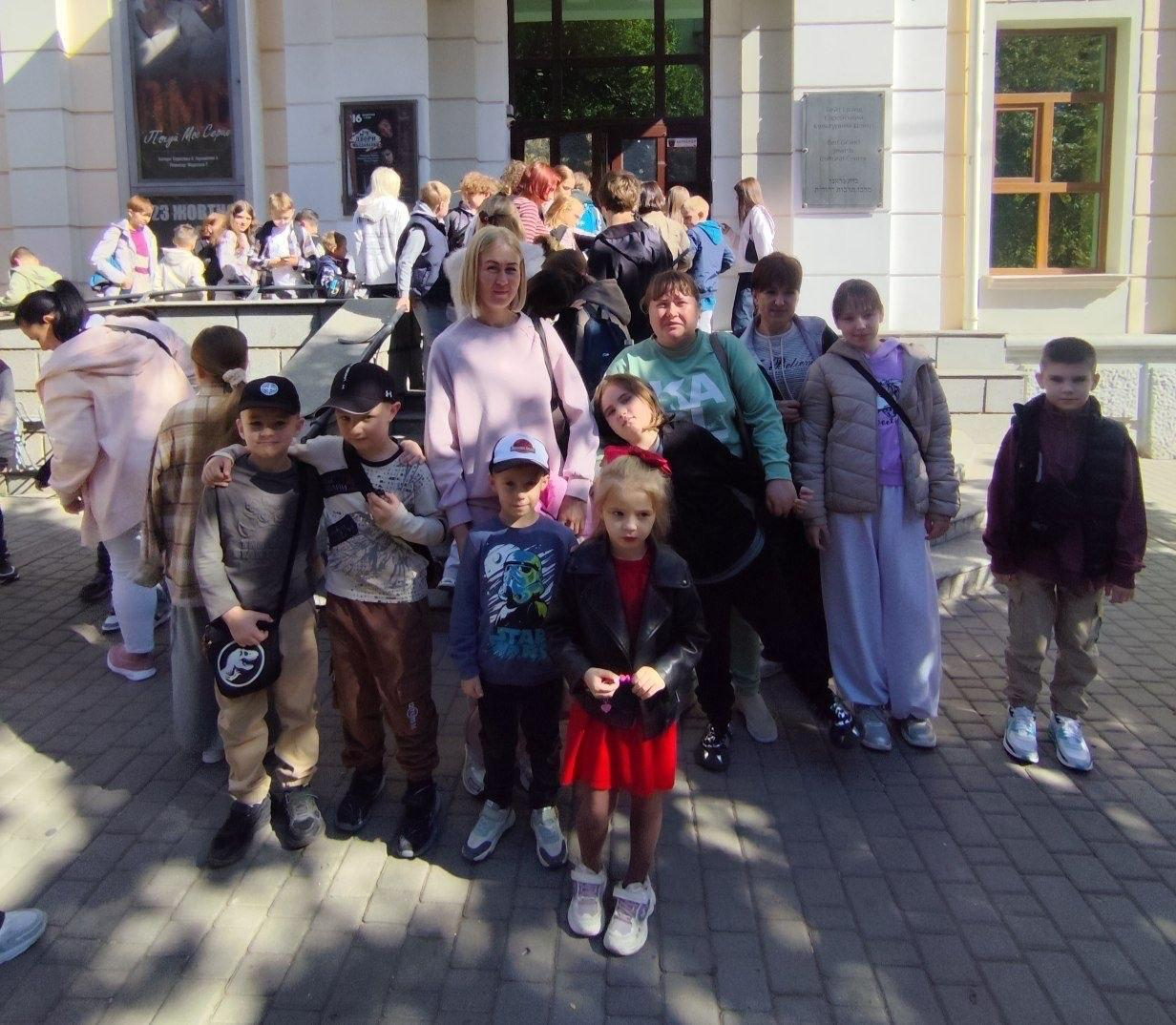Група маленьких переселенців із Херсонщини разом із мамами позує для спільного пам'ятного фото на вимощеному бруківкою подвір'ї перед входом до Єврейського культурного центру «Beit Grand» в Одесі після відвідування дитячої театральної вистави «Мауглі». На передньому плані кадру стоять діти у повсякденному одязі та бейсболках, серед яких хлопчик у сірому світшоті з круглою чорною сумкою через плече, маленька дівчинка у яскраво-червоній сукні та чорній шкіряній куртці-косусі, а також хлопчик у синьому лонгсліві із зображенням героїв Зоряних війн. Позаду дітей розмістилися мами та супроводжуючі жінки, зокрема блондинка у світло-рожевому светрі та її колега у бірюзовому світшоті з написом, а на задньому плані біля сходів та скляних вхідних дверей будівлі помітно велику кількість інших юних глядачів та розвішані на стінах театральні афіші з репертуаром центру. Культурний захід для психоемоційного розвантаження сімей ВПО організовано координаційним центром «Вільні разом» спільно з БФ «Сапорт Херсон» та за сприяння Департаменту соціального розвитку Херсонської ОДА.