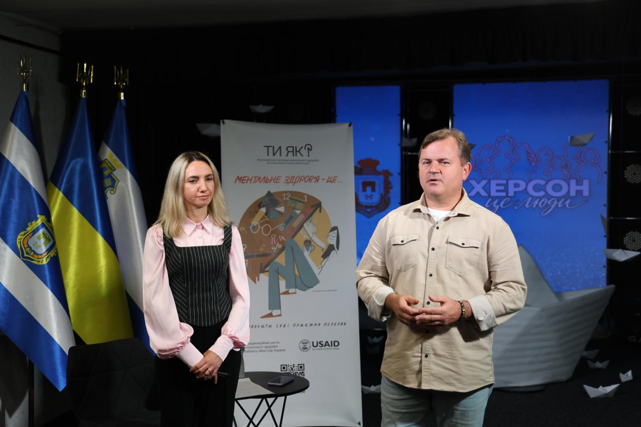Заступник начальника Херсонської ОДА Володимир Клюцевський та регіональна координаторка Всеукраїнської програми ментального здоров'я «Ти як?» у Херсонській області Ілона Задорожня виступають перед присутніми під час відкриття Дня спільнодії до Дня ментального здоров’я у Херсоні. Праворуч у кадрі Володимир Клюцевський у бежевій сорочці та світлих джинсах активно жестикулює під час промови, наголошуючи на важливості розвитку доступних психосоціальних послуг в умовах війни, водночас ліворуч від нього уважно слухає колегу Ілона Задорожня у світло-рожевій блузці й чорному жилеті. Між промовцями на передньому плані встановлено невеликий круглий столик із мобільним телефоном, за яким височіє великий вертикальний інформаційний банер програми «Ти як?» із написом «МЕНТАЛЬНЕ ЗДОРОВ'Я — ЦЕ...» та емблемою USAID у нижній частині. Робоча зустріч для підтримки благополуччя мешканців громад проходить у спеціально облаштованій залі, де ліворуч на флагштоках піднято прапор міста Херсона, Державний прапор України та прапор Херсонської області, а на задньому плані по центру встановлено синій тематичний екран із офіційним гербом м. Херсона та великим художнім написом «ХЕРСОН це люди».