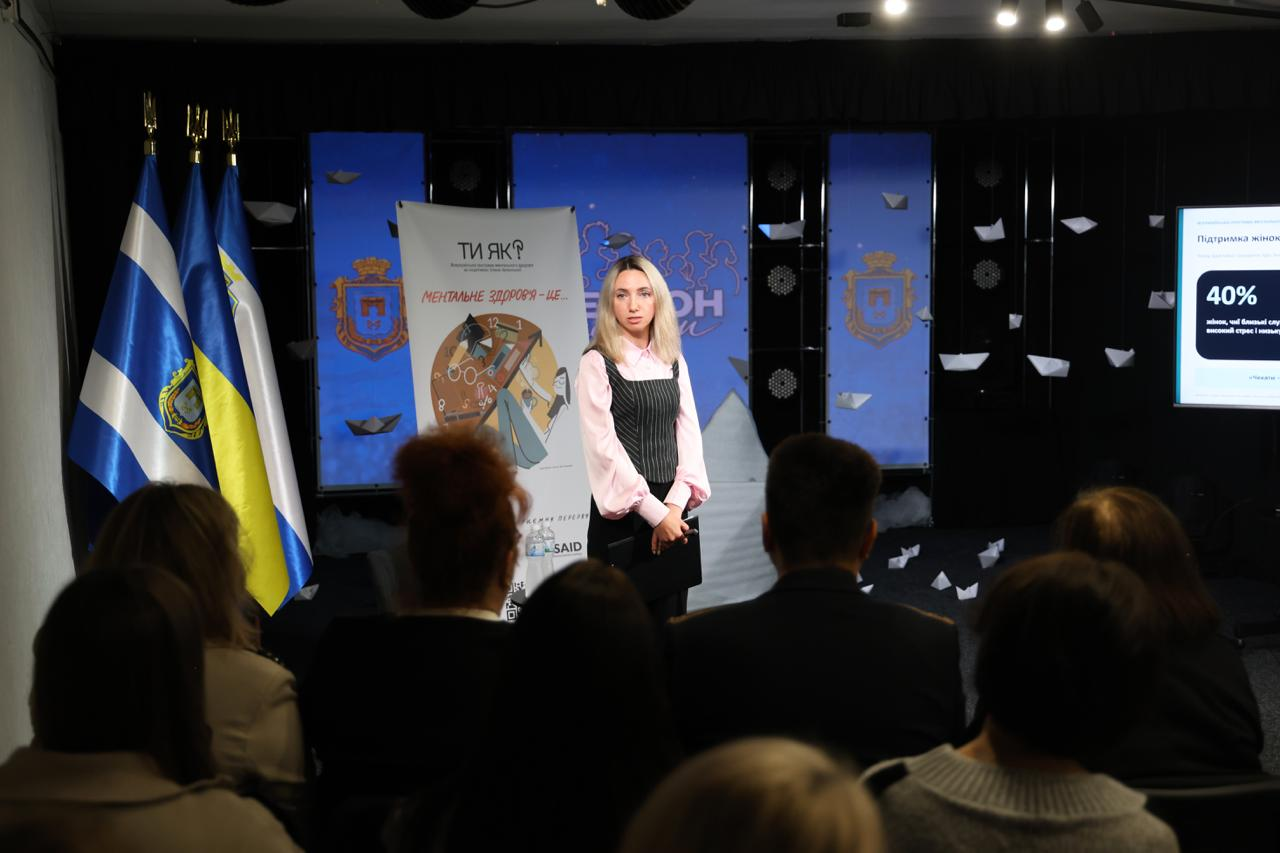 Регіональна координаторка Всеукраїнської програми ментального здоров'я «Ти як?» у Херсонській області Ілона Задорожня виступає на сцені перед слухачами під час презентації комунікаційної кампанії в межах Дня спільнодії до Дня ментального здоров’я у Херсоні. Координаторка у світло-рожевій блузці та чорному жилеті стоїть по центру сцени зі складеними руками та тримає папку з матеріалами доповіді, розповідаючи присутнім у залі про нову хвилю кампанії «Підтримка жінок, чиї близькі служать» та її головний меседж «Чекати – це також дія». На передньому плані спиною до об'єктива сидить велика аудиторія фахівців та представників громад, які уважно слухають спікерку, а ліворуч від неї на сцені встановлено великий вертикальний інформаційний банер програми «Ти як?» із написом «МЕНТАЛЬНЕ ЗДОРОВ'Я — ЦЕ...» та емблемою USAID. Урочистий та просвітницький захід проходить у залі із чорним покриттям підлоги, де ліворуч на дерев'яних флагштоках піднято прапор міста Херсона, Державний прапор України та прапор Херсонської області, по центру на задньому плані змонтовано синій екран із офіційним гербом м. Херсона, художнім неоновим написом «ХЕРСОН це люди» та інсталяцією з декоративних білих паперових корабликів, а праворуч розташовано великий білий мультимедійний екран із важливою статистикою про «40%» жінок, чиї близькі перебувають на службі у війську.