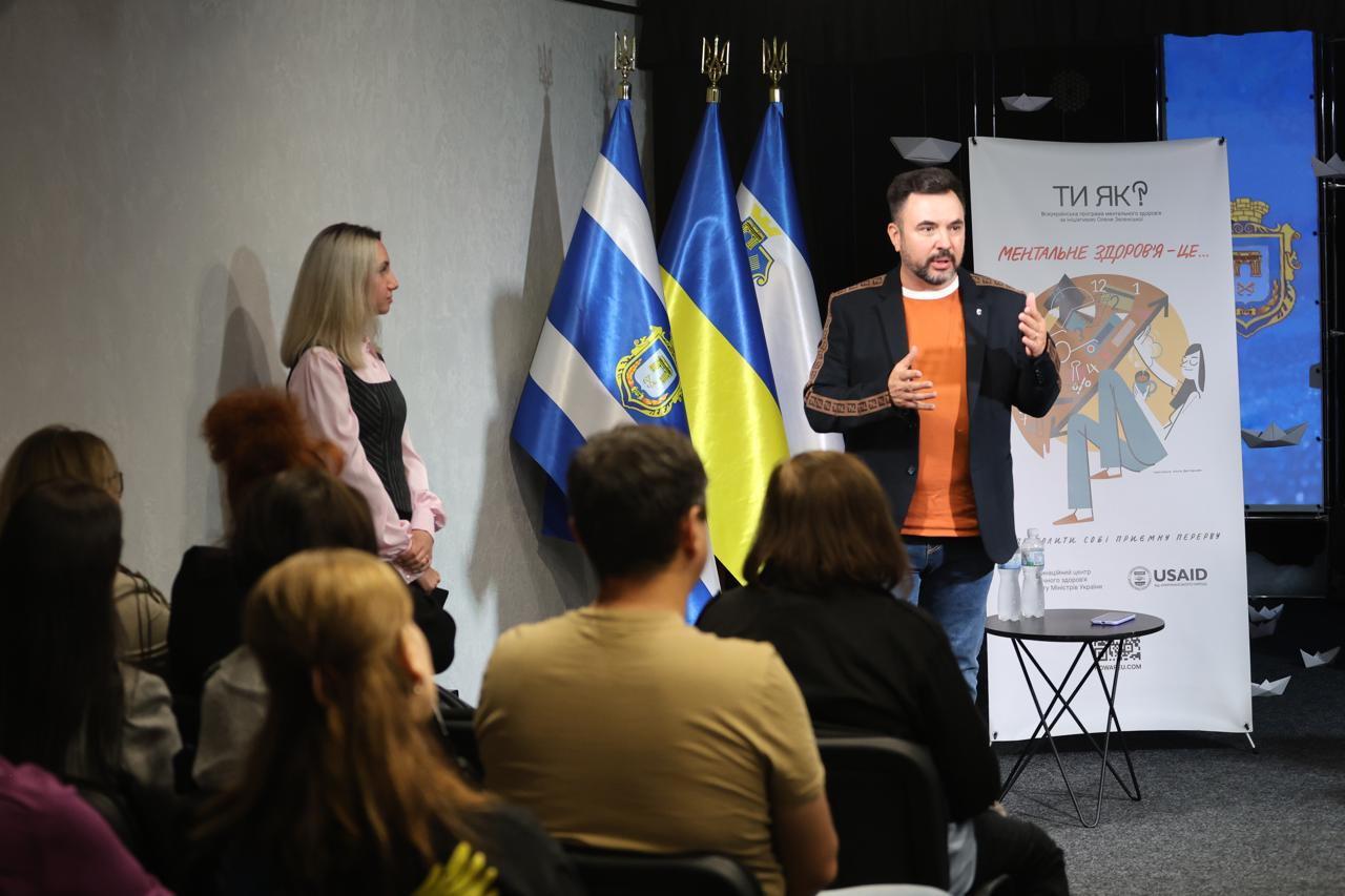 Заступник начальника Департаменту реалізації гуманітарної політики — начальник управління культури Херсонської ОДА Андрій Ярьоменко виступає на сцені під час Дня спільнодії до Дня ментального здоров’я у Херсоні. Спікер у чорному піджаку, помаранчевій футболці та синіх джинсах активно жестикулює під час промови, ділячись практичним досвідом реалізації регіонального проєкту «Мистецтво відновлення та підтримки мешканців громад» та роллю закладів культури як просторів психосоціальної підтримки, а безпосередньо праворуч від Андрія Ярьоменка знаходиться маленький круглий стіл із пляшками води. Ліворуч у кадрі уважно слухає виступ регіональна координаторка Всеукраїнської програми ментального здоров'я «Ти як?» у Херсонській області Ілона Задорожня у світло-рожевій блузці й чорному жилеті, а на передньому плані спиною до об'єктива сидить велика аудиторія слухачів та експертів прифронтових громад. За спиною Андрія Ярьоменка височіє великий вертикальний інформаційний банер програми «Ти як?» із написом «МЕНТАЛЬНЕ ЗДОРОВ'Я — ЦЕ...» та емблемою USAID у нижній частині. Робоча зустріч проходить у залі, де ліворуч на флагштоках піднято прапор міста Херсона, Державний прапор України та прапор Херсонської області, а на задньому плані сцени змонтовано синій тематичний екран із офіційним гербом м. Херсона.