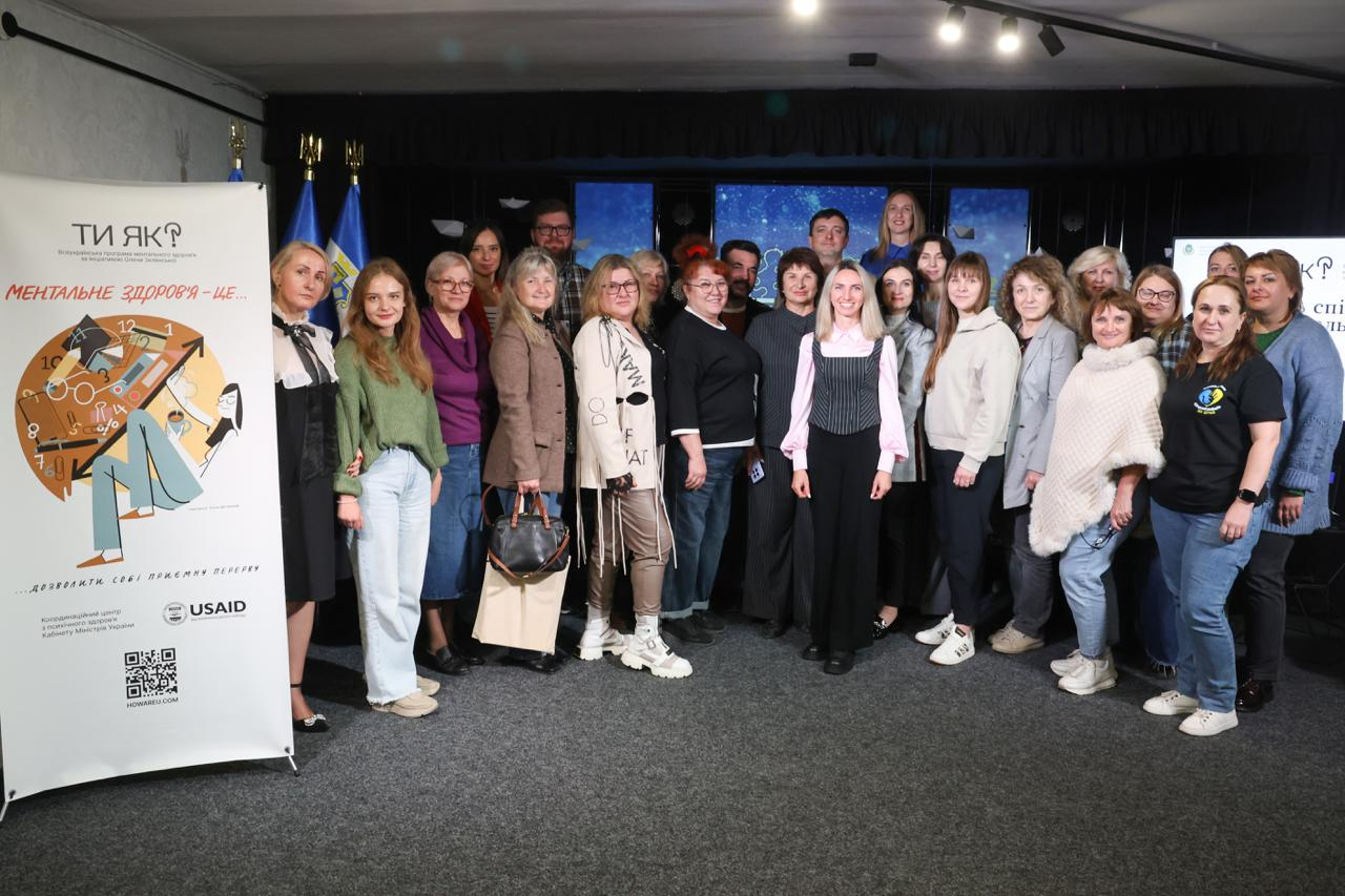 Велика група учасників заходу, представників влади, психологів та координаторів громад позує для спільного пам'ятного фото на сцені під час завершення Дня спільнодії до Дня ментального здоров’я у Херсоні. Численні експерти Всеукраїнської програми ментального здоров'я «Ти як?» вишикувалися у два ряди по всій ширині сцени, посміхаючись в об'єктив кадру та демонструючи єдність у підтримці психоемоційного благополуччя мешканців регіону. З лівого краю у першому ряду стоять учасниця у чорній кофті з білим коміром, дівчина у зеленому светрі та світлих джинсах, а також жінка у фіолетовій кофті та жінка у коричневому кардигані з великою сумкою в руках. У самому центрі першого ряду перебуває регіональна координаторка програми «Ти як?» у Херсонській області Ілона Задорожня у світло-рожевій блузці й чорному жилеті, безпосередньо за лівим плечем якої у другому ряду стоїть заступниця Херсонського міського голови з питань діяльності виконавчих органів ради Наталія Чорненька, а ліворуч від Наталії Чорненької у тому ж ряду перебуває заступник начальника Департаменту реалізації гуманітарної політики — начальник управління культури Херсонської ОДА Андрій Ярьоменко. Праворуч від координаторки у першому ряду розмістилися жінки у світлих кофтах, сірих піджаках та колега у чорній футболці з офіційним круглим логотипом програми «Ти як?». З лівого боку сцени поруч із першим рядом виставлено великий вертикальний інформаційний банер із написом «МЕНТАЛЬНЕ ЗДОРОВ'Я — ЦЕ...» та емблемою USAID, трохи далі за ним на флагштоках піднято прапор міста Херсона, Державний прапор України та прапор Херсонської області, а вся урочиста зустріч проходить на тлі синього центрального екрана з офіційним гербом м. Херсона.