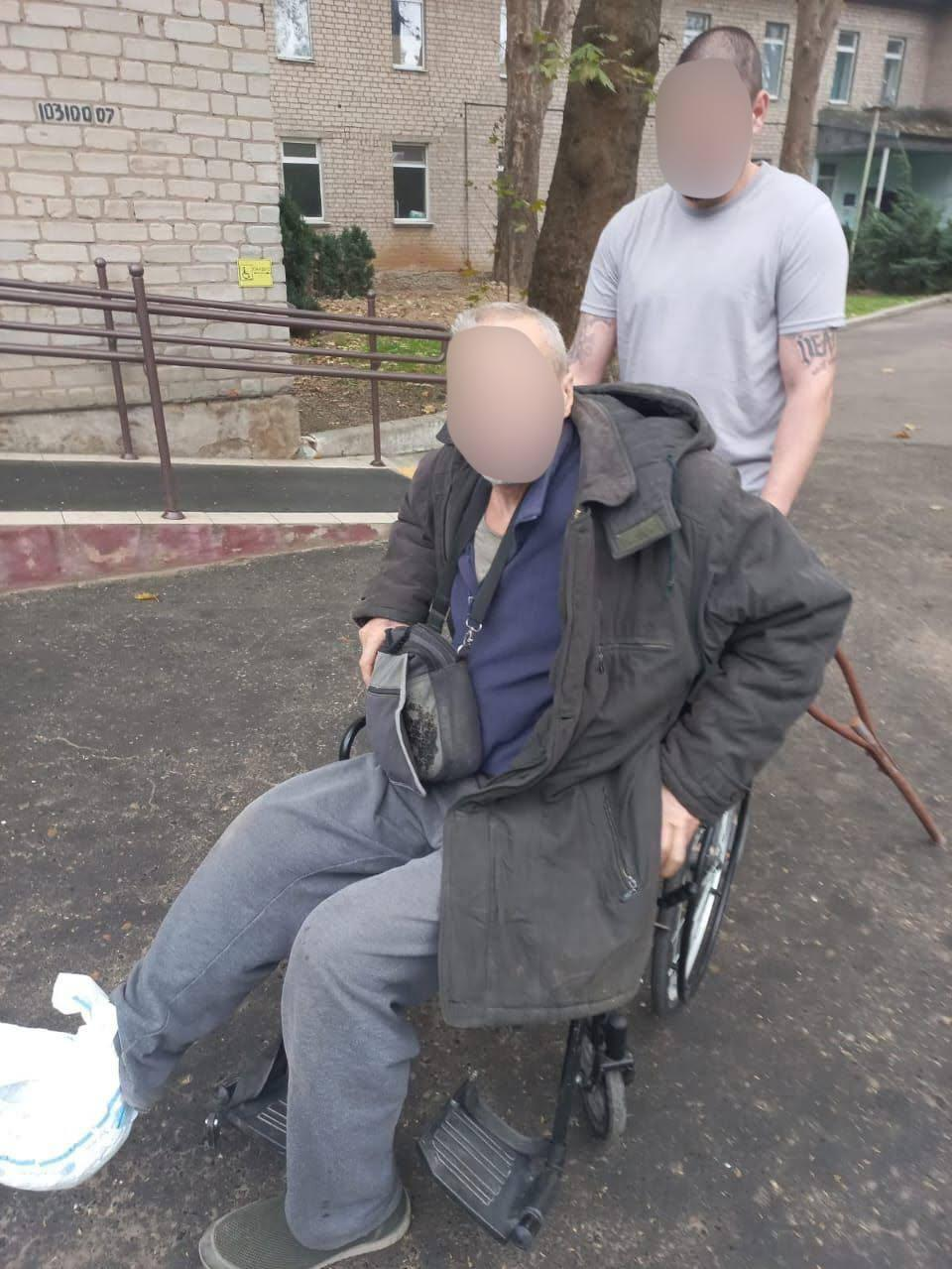 Український військовослужбовець першого батальйону 102-ї окремої бригади територіальної оборони супроводжує евакуйованого з фронтового Берислава пораненого місцевого жителя на інвалідному візку біля медичного закладу. На передньому плані ліворуч у кріслі-колісному сидить літній чоловік у подовженій темній зимовій куртці з капюшоном, синій сорочці поло, сірих спортивних штанях та капцях, який тримає на колінах невелику чорну сумку через плече, а ліворуч біля його ніг стоїть білий пакет із речами. Безпосередньо за його спиною стоїть міцний військовий у сірій футболці з татуюваннями на руках, який дбайливо тримає ручки візка, допомагаючи пораненому, що тримає дерев'яну опорну палицю з правого боку. Процес успішного вивезення громадянина з-під російських ударів та передачі його лікарям зафіксовано на свіжому асфальтованому майданчику на тлі світлої цегляної будівлі лікарні, де ліворуч облаштовано спеціальний пандус із металевими поручнями для маломобільних груп населення, а на задньому плані видно зелені дерева та вікна палат.