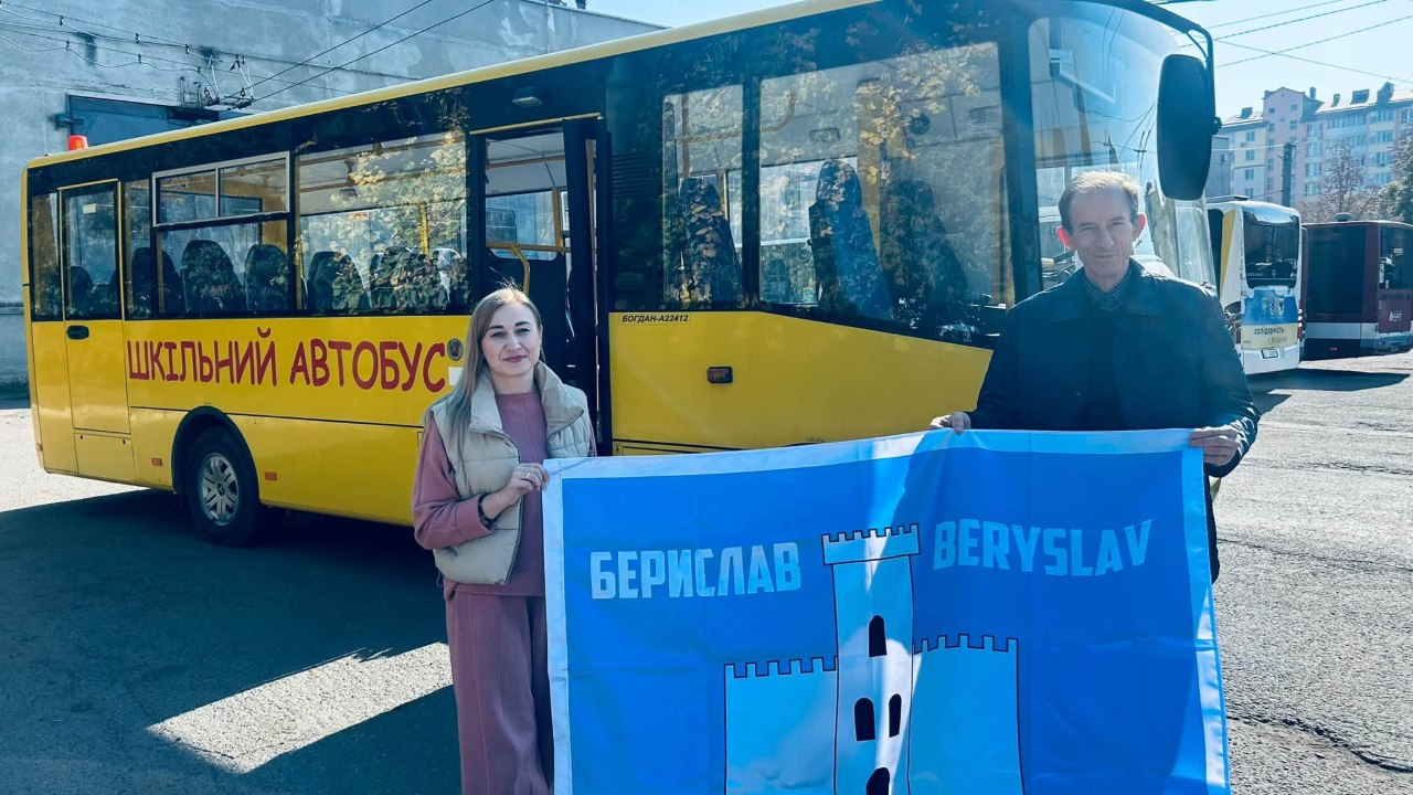 Жінка та чоловік спільно тримають великий прапор міста Берислав перед яскраво-жовтим пасажирським автобусом, який було передано для підвезення школярів. Ліворуч у кадрі стоїть молода жінка з довгим світлим волоссям у рожевому костюмі та світлій дутій жилетці, яка посміхається та тримає край синього полотнища з білим зображенням фортечної вежі та офіційними написами «БЕРИСЛАВ» і «BERYSLAV». Праворуч від неї стоїть чоловік у темній куртці, який підтримує інший бік міського стяга. Безпосередньо за їхніми спинами розташований пасажирський автобус марки «Богдан» із відкритими передніми дверима та великим червоним написом «ШКІЛЬНИЙ АВТОБУС» на борту під вікнами. Передача транспорту для релокованих закладів освіти зафіксована у світлу сонячну погоду на асфальтованій території на тлі міських будівель, а на задньому плані праворуч помітно інші автобуси та тролейбусні лінії.
