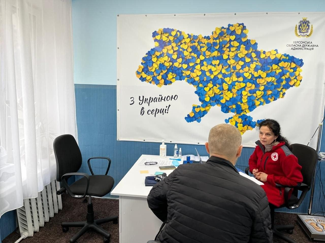 Мобільна бригада Товариства Червоного Хреста України забезпечила необхідну медичну підтримку для внутрішньо переміщених осіб у приміщенні херсонського гуманітарного центру «Вільні разом» у Хмельницькому, де під час прийому дев’ятеро людей отримали фахові консультації та лікарські засоби. На фотографії зафіксовано момент індивідуального прийому, під час якого темноволоса фахівчиня у фірмовій яскраво-червоній куртці з білою круглою емблемою організації сидить праворуч за білим робочим столом і веде діалог із відвідувачем у чорній дутій куртці, що розташований спиною до об'єктива. На поверхні столу між ними розкладено медичні документи, бланки, антисептичні засоби та відкритий ноутбук для фіксації даних, а ліворуч біля великого вікна з білим тюлем та батареєю опалення облаштовано додаткове робоче місце з вільним чорним кріслом. Зустріч проходить у затишній світлій кімнаті з синіми стінами та темним ковроліном на тлі великого настінного білого банера, прикрашеного патріотичним гербом Херсонської обласної державної адміністрації, художнім написом «З Україною в серці!» та великою картою України, яка повністю викладена з маленьких жовтих і блакитних сердечок.