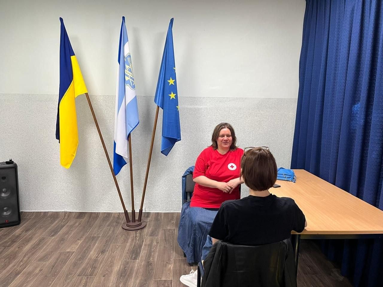 Психологиня Товариства Червоного Хреста України проводить індивідуальну консультацію для відвідувачки в кабінеті херсонського гуманітарного центру «Вільні разом» у Хмельницькому, де протягом тижня одинадцятеро людей отримали необхідну психологічну допомогу та підтримку. У центрі кадру за довгим дерев'яним робочим столом сидить психологиня організації у фірмовій яскраво-червоній футболці з круглою білою емблемою та довгій синій спідниці, яка доброзичливо посміхається та спілкується з жінкою, що сидить перед нею спиною до об'єктива у чорній футболці. На столі праворуч лежить невелика яскраво-блакитна жіноча сумочка та мобільний телефон, а з лівого боку кабінету на підлозі встановлено чорну музичну або комп'ютерну колонку. Індивідуальна зустріч для емоційного розвантаження та підтримання ментального здоров'я внутрішньо переміщених осіб проходить у світлій кімнаті з сіро-білими стінами та лінолеумом під дерево на тлі великої синьої захисної штори праворуч та трьох піднятих на спільній дерев'яній підставці прапорів ліворуч — Державного прапора України, біло-блакитного прапора Херсонської області та синього прапора Європейського Союзу з жовтими зірками.