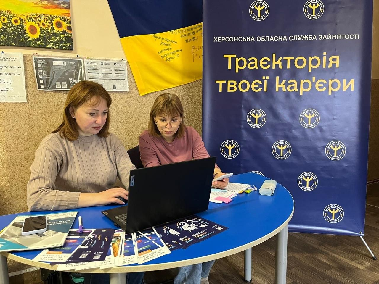 Працівниця центру зайнятості надає індивідуальну консультацію з питань працевлаштування та кар'єрного розвитку жінці-переселенці у приміщенні херсонського гуманітарного центру «Вільні разом» у Хмельницькому. Ліворуч за круглим синім столом сидить русява фахівчиня у сірому джемпері, яка зосереджено працює за відкритим чорним ноутбуком, водночас поруч із нею сидить відвідувачка у рожевій кофті, яка уважно вивчає інформацію на екрані комп'ютера. На передньому плані столу розкладено велику кількість офіційних друкованих інформаційних буклетів, брошур та листівок Державної служби зайнятості із заголовками про можливості для молоді, а поруч лежать мобільні телефони, канцелярія та кольорові папірці для нотаток. Індивідуальний прийом громадян для всебічної підтримки внутрішньо переміщених осіб проходить у робочому кабінеті на тлі світло-жовтої стіни, де ліворуч висять інформаційні плакати та підписаний маркерами Державний прапор України, а праворуч встановлено великий вертикальний темно-синій банер із численними круглими емблемами, офіційним написом «ХЕРСОНСЬКА ОБЛАСНА СЛУЖБА ЗАЙНЯТОСТІ» та великим жовто-білим заголовком «Траєкторія твоєї кар'єри».