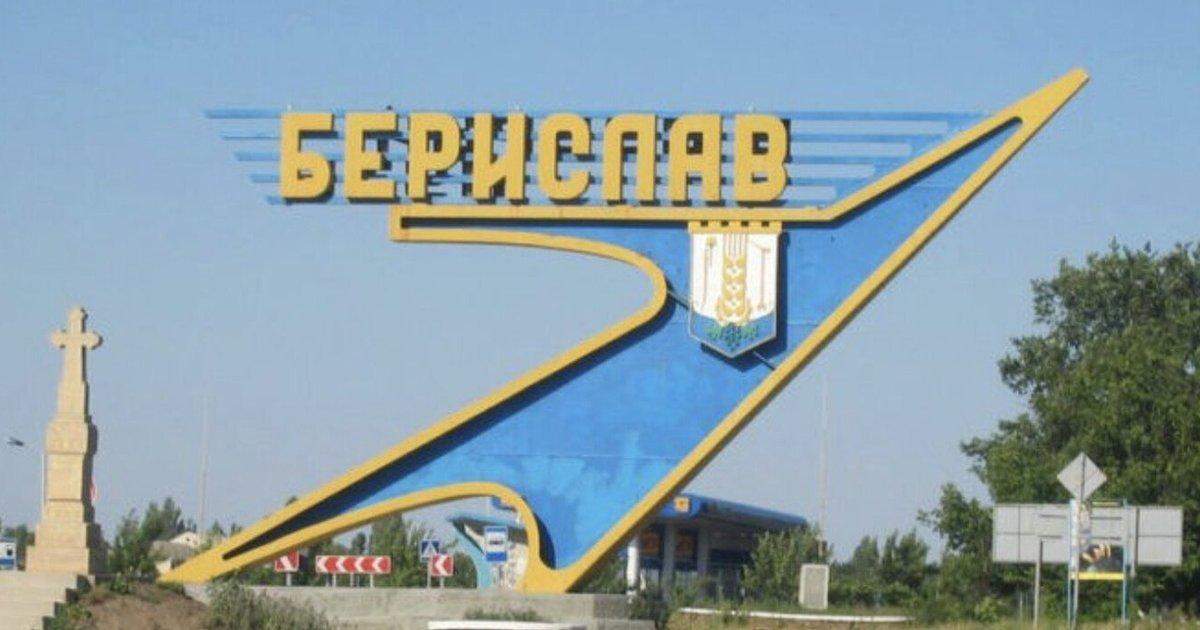 Великий синьо-жовтий архітектурний знак-стела з об'ємними жовтими літерами «БЕРИСЛАВ» та офіційним міським гербом встановлений біля автомобільної дороги під безхмарним блакитним небом фронтового міста. До цього прифронтового населеного пункту на лінії вогню продовжуються гуманітарні місії: цієї ночі бійці підрозділу «Болгаричі» 11-ї бригади Нацгвардії за допомогою дронів доставили мешканцям ще 460 кілограмів життєво необхідного продовольства. На самому знімку ліворуч від знака височіє кам'яний пам'ятник у вигляді хреста на постаменті, а на задньому плані вздовж автошляху помітно елементи інфраструктури, дорожні знаки та зелені дерева.