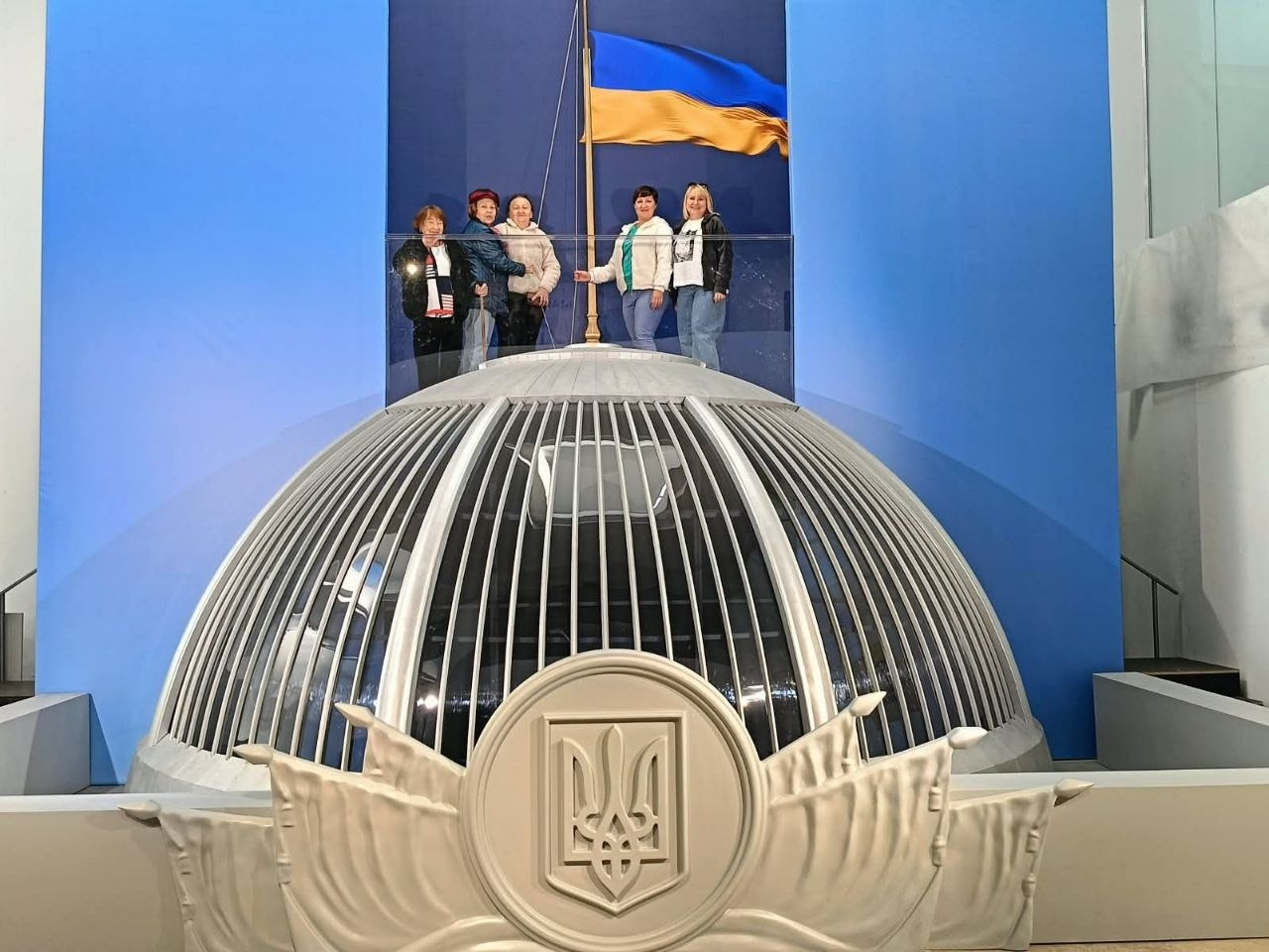 Група жінок-переселенок із Херсонщини позує для пам'ятного фото на оглядовому майданчику інтерактивної виставки «Той день» у Львові, символічно піднявши Державний прапор України над реалістичною масштабною інсталяцією купола Верховної Ради. Учасниці екскурсії, організованої координаційним центром «Вільні разом», стоять у ряд на височині за прозорим скляним огородженням безпосередньо біля високого дерев'яного флагштока з майоріючим синьо-жовтим стягом. Жінки різного віку вбрані у повсякденні куртки, кофти та джинси, а вся виставкова зона оформлена у насичених яскраво-блакитних кольорах. На передньому плані кадру крупно представлений масивний світлий купол будівлі парламенту з металевими вертикальними ребрами-ґратами, прикрашений по центру великим рельєфним зображенням офіційного Державного Герба України — Тризуба, під яким висічено декоративні елементи у вигляді розгорнутих знамен.