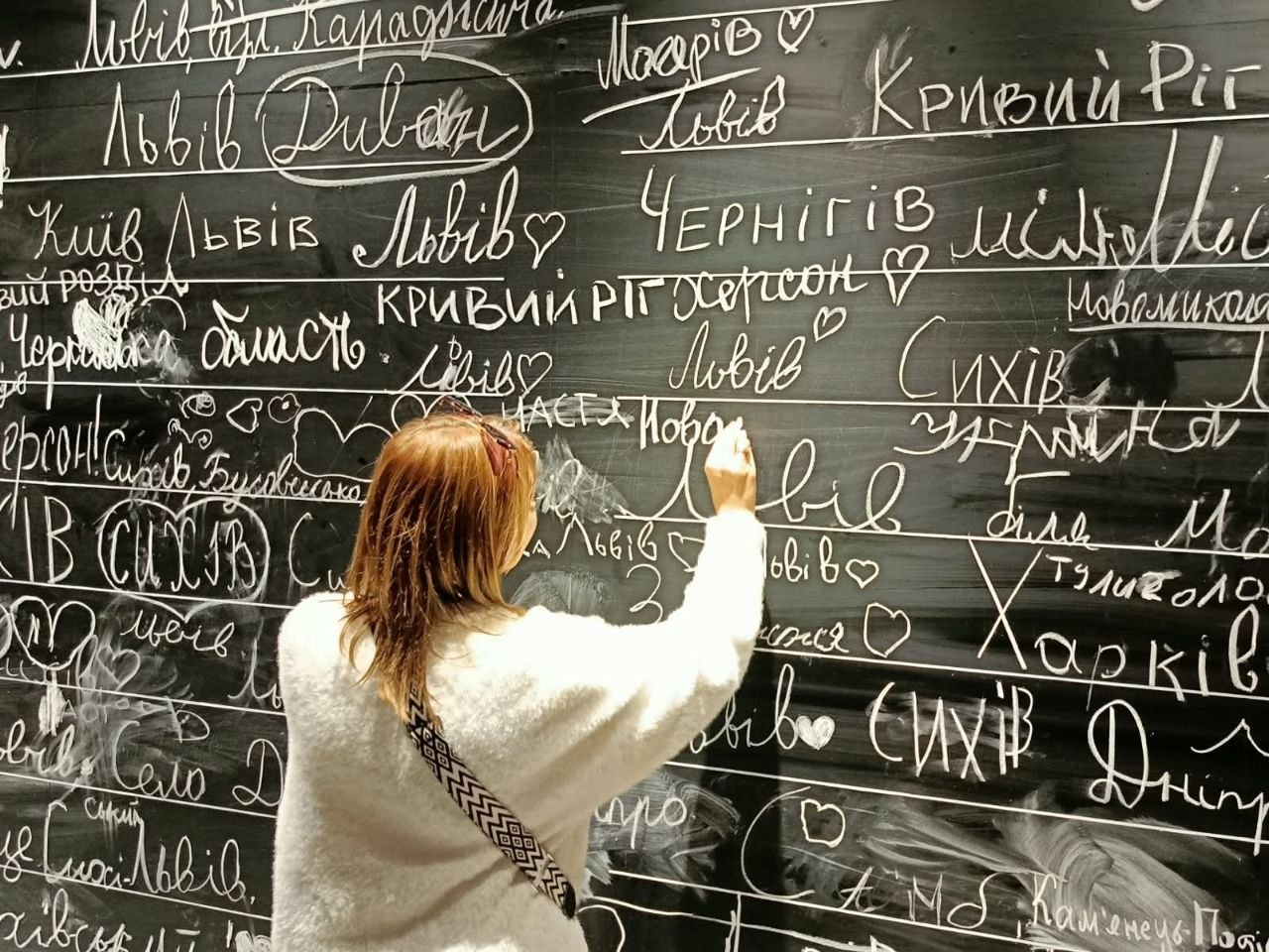 Учасниця екскурсії для внутрішньо переміщених осіб із Херсонщини залишає свій запис крейдою в «зоні мрій» під час відвідування інтерактивної виставки «Той день» у Львові. Жінка з русявим волоссям у світлій пухнастій кофті та з чорно-білою сумкою на тканинному ремені через плече стоїть спиною до об'єктива по центру кадру й виводить напис на великій чорній грифельній стіні-дошці. Уся поверхня інтерактивної стіни густо заповнена численними написами та назвами міст, які білою крейдою залишили інші відвідувачі, де чітко видно слова «Херсон», «Кривий Ріг», «Чернігів», «Харків», «Дніпро», «Київ», «Львів», «Сихів» та намальовані контури сердечок, відображаючи сподівання сімей ВПО про майбутній день перемоги України та повернення до рідних домівок.