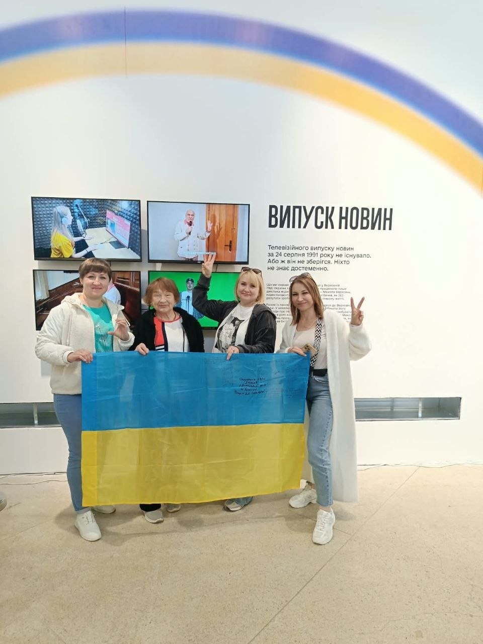 Учасниці екскурсії для внутрішньо переміщених осіб із Херсонщини розгортають Державний прапор України під час відвідування інтерактивної виставки «Той день» у Львові. Жінки різного віку позують для пам'ятного фото у світлій залі координаційного центру біля інтерактивного медіастенда з великим чорним написом «ВИПУСК НОВИН» та історичним текстом про події 24 серпня 1991 року. Присутні вбрані у світлі кофти, футболки, джинси та довгий білий кардиган, посміхаються в об'єктив кадру та показують жести єдності й перемоги пальцями рук, тримаючи перед собою велике синьо-жовте полотнище з підписами. На стіні над головами екскурсанток облаштовано мультимедійну зону з чотирма увімкненими моніторами, які демонструють кадри з телевізійної студії та засідань, а вгорі інтер'єр доповнений великою плавною дугою у патріотичних кольорах прапора.