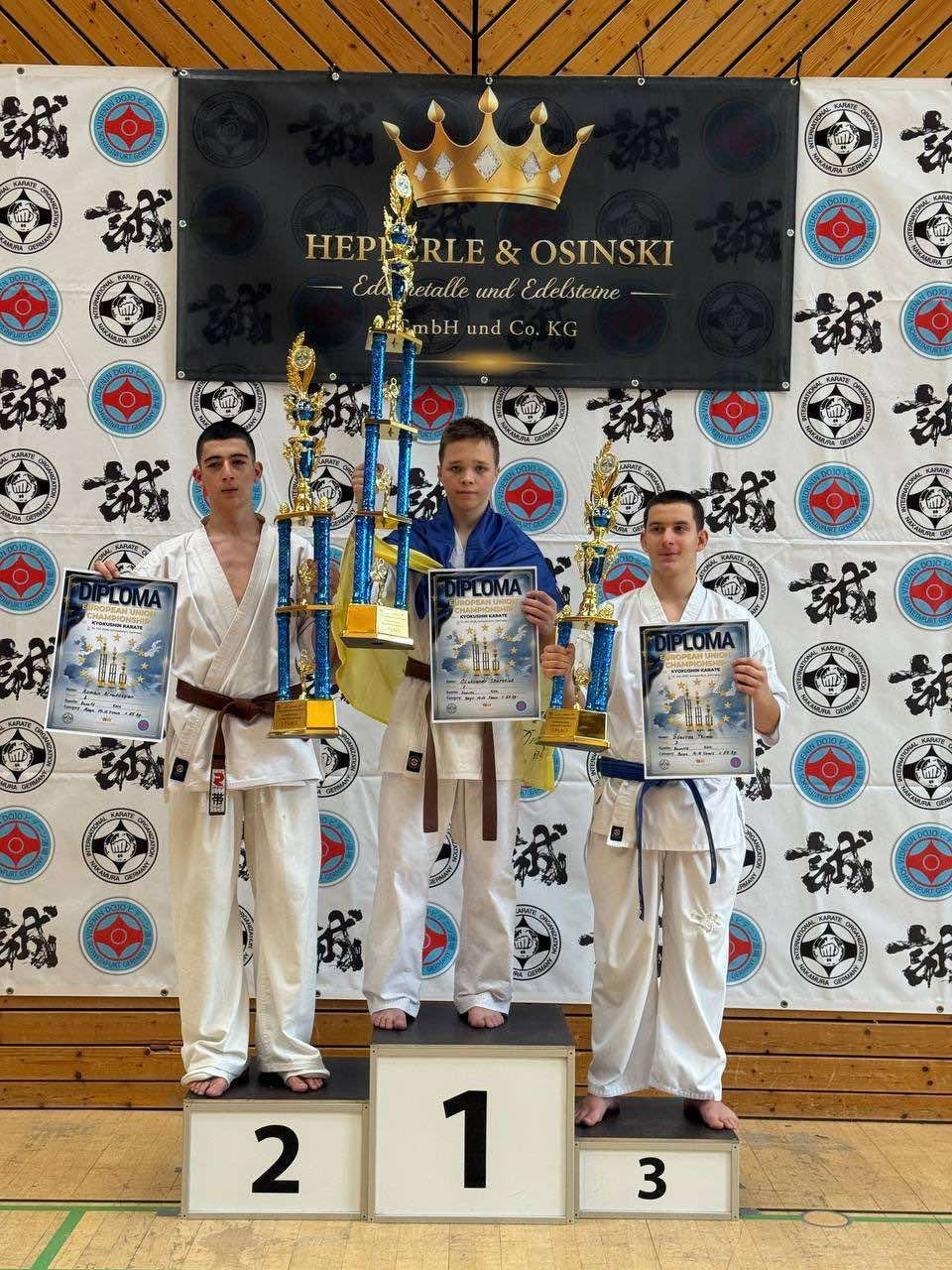 Три юні українські каратисти-призери Чемпіонату Європи з кіокушин БуДо карате за версією IKON урочисто позують на спортивному п'єдесталі пошани у німецькому місті Швайнфурт. Усі спортсмени вбрані у білосніжні кімоно-догі з фірмовими нашивками та тримають у руках великі золотисті нагородні кубки з блакитними елементами та офіційні дипломи чемпіонату «DIPLOMA European Union Championships Kyokushin Karate». По центру на найвищій сходинці під номером один стоїть переможець Олександр Шерстюк із коричневим поясом, плечі якого дбайливо оповиті синьо-жовтим Державним прапором України. Ліворуч від нього на другій позиції розмістився срібний призер із коричневим поясом, а праворуч на третій сходинці стоїть босоногий каратист із синім поясом. Урочиста церемонія нагородження вихованців дитячо-юнацької школи проходить на тлі великого світлого тематичного банера з численними круглими емблемами та японськими ієрогліфами-канManagement, над якими по центру закріплено темний плакат спонсорів «HEPPERLE & OSINSKI Edelmetalle und Edelsteine» з великим золотим малюнком корони.