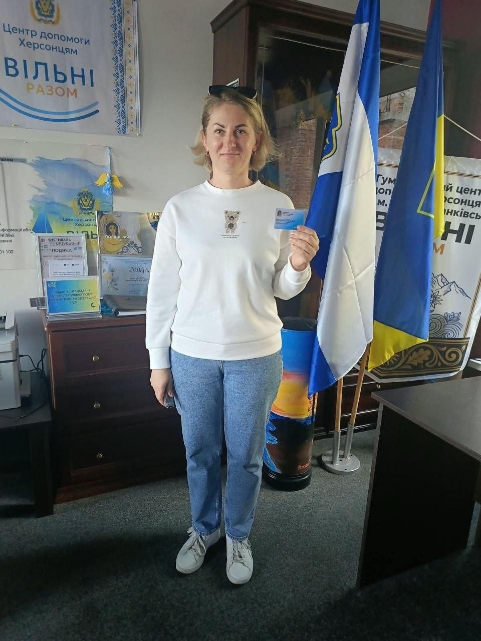 Жінка-переселенка з Херсонщини тримає в руці отриману пластикову картку, позуючи в офісі координаційного центру «Вільні разом» в Івано-Франківську в межах реалізації проєкту «Картка Херсонщини». Учасниця зустрічі у світлому світшоті з невеликим принтом ведмедика, синіх джинсах та білих кросівках посміхається в об'єктив кадру, демонструючи нову іменну картку для підтримки ВПО. Вона стоїть по центру кімнати на тлі офіційних символів, де праворуч на підставках встановлено Державний прапор України та біло-блакитний прапор Херсонської області, а ліворуч на комоді розміщено численні нагородні дипломи, подяки та великий настінний банер із логотипом «Центр допомоги херсонцям ВІЛЬНІ РАЗОМ».