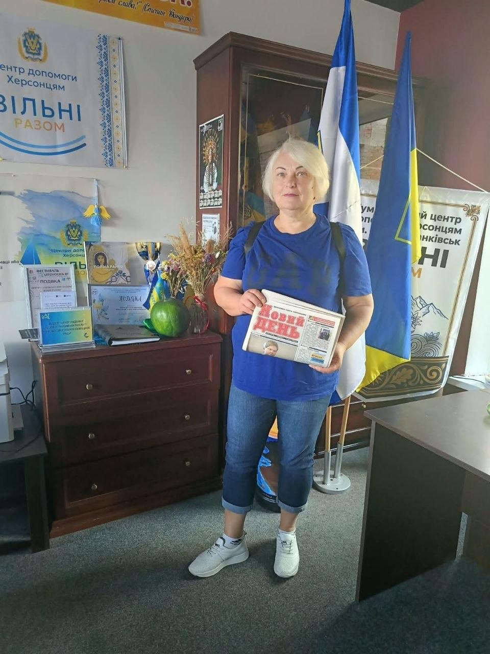 Жінка-переселенка з Херсонщини тримає в руках свіжий примірник газети, позуючи в офісі координаційного центру «Вільні разом» в Івано-Франківську під час отримання українських друкованих видань. Учасниця зустрічі зі світлим волоссям, вбрана в яскраво-синю футболку з патріотичним принтом, сині джинси та білі кросівки, демонструє номер газети «Новий день» із червоним заголовком. Вона стоїть по центру робочого кабінету, де праворуч на підставках піднято Державний прапор України та біло-блакитний прапор Херсонської області, а ліворуч на дерев'яному комоді облаштовано тематичний куточок із дипломами, фестивальними подяками, мапою України та фірмовим настінним банером «Центр допомоги херсонцям ВІЛЬНІ РАЗОМ».