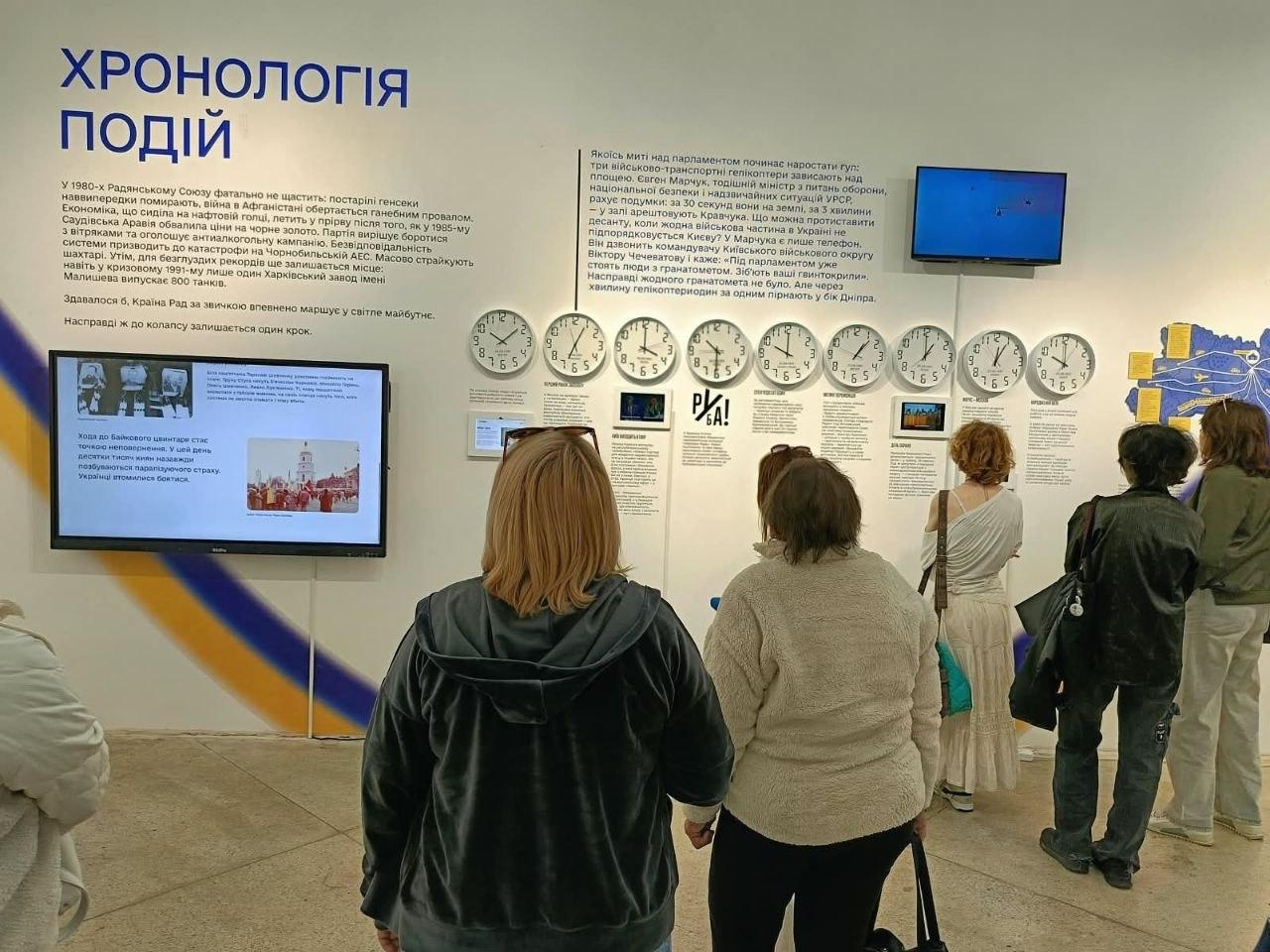 Жінки-переселенки з Херсонщини уважно вивчають інформаційні стенди інтерактивної виставки «Той день» під час подорожі до Львова, присвяченої історії становлення української державності. Учасниці екскурсії, організованої координаційним центром «Вільні разом», стоять спиною до об'єктива кадру у виставковій залі перед великою світлою стіною з експозицією під назвою «ХРОНОЛОГІЯ ПОДІЙ». На стіні розміщено великі текстові блоки про історичні події кінця 1980-х та початку 1991 року, розвішано довгий ряд круглих настінних годинників-циферблатів та встановлено невеликі мультимедійні екрани, що транслюють архівні відеоматеріали. Жінки вбрані у повсякденний осінній одяг, зокрема у темні кофти, куртки та світлі штани, а з лівого боку біля них нахилився інформаційний монітор із презентаційним слайдом про ядерне роззброєння та страхи епохи, допомагаючи родинам ВПО глибше зануритися в атмосферу знакових днів проголошення незалежності України.