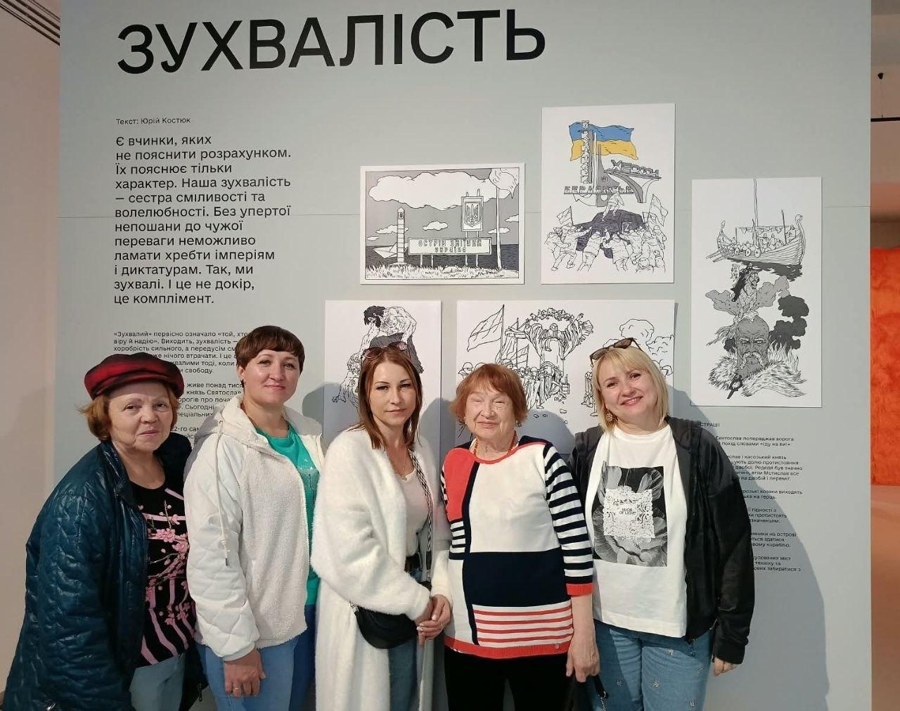 Група жінок-переселенок із Херсонщини позує для пам'ятної світлини у виставковій залі інтерактивної виставки «Той день» під час екскурсійної поїздки до Львова, присвяченої подіям становлення української державності. Учасниці заходу, організованого координаційним центром «Вільні разом», стоять у ряд, посміхаючись в об'єктив кадру на тлі великого світлого тематичного стенда з великим капітальним написом «ЗУХВАЛІСТЬ». На стіні за їхніми спинами розміщено офіційні текстові матеріали про національний характер, сміливість та боротьбу українців, а також закріплено оригінальні патріотичні малюнки й карикатури, серед яких чітко виділяється ескіз прикордонного стовпа з написом «Острів Зміїний». Жінки різного віку вбрані у різнокольоровий повсякденний осінній одяг, зокрема у темні кофти з візерунками, світлі светри, білу футболку та бірюзову блузку, а учасниця з лівого краю додатково одягла класичний червоний берет, що додає подорожі родин ВПО особливої атмосфери єдності та культурного відродження.