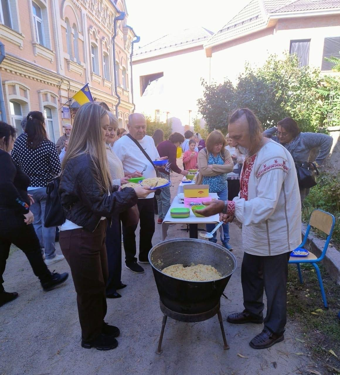 На подвір'ї кропивницького координаційного центру «Вільні разом», який урочисто відзначив другу річницю з моменту заснування, організували традиційне частування козацьким кулешем для переселенців із Херсонщини. На світлині літній кухар у білій вишиванці з автентичним червоно-чорним орнаментом насипає ложкою гарячу страву з великого чорного чавунного казана на залізній підставці. З лівого боку біля вогню вишикувалася черга з гостей заходу, де на передньому плані молода дівчина у чорній шкіряній куртці тримає тарілку, чекаючи на частування, а поруч облаштовано довгий стіл із білою скатертиною та кольоровим одноразовим посудом. Святкова гуманітарна акція для всебічної підтримки сімей ВПО проходить у ясну сонячну погоду біля гарної цегляної будівлі хабу, над входом до якої гордо майорить Державний прапор України.