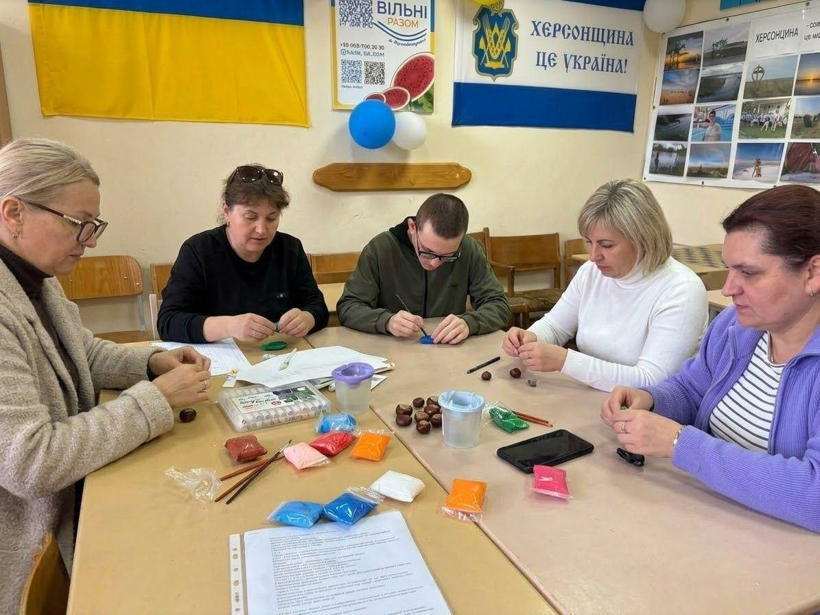Переселенці з Херсонщини зосереджено створюють святкові поробки своїми руками, сидячи за великим спільним столом в офісі координаційного центру «Вільні разом» у Кропивницькому під час творчого майстер-класу, який спільно з благодійним фондом «Медицина Херсонщини» присвятили Міжнародному дню вчителя. Учасники заходу — жінки та юнак — уважно працюють з матеріалами, розробляючи елементи для майбутніх квіткових композицій. На столі перед ними розкладено велику кількість яскравих різнокольорових пакетиків з повітряними кульками, каштани для декору, пластикові ємності, пензлики та роздруковані інформаційні інструкції. Зустріч для психоемоційного розвантаження та підтримки родин ВПО проходить у затишному патріотичному кабінеті хабу, де стіни прикрашені Державним прапором України, великим біло-синім банером із золотим тризубом та офіційним написом «Херсонщина — це Україна!», а також інформаційним плакатом з QR-кодом та колажем із мальовничими краєвидами рідного краю.