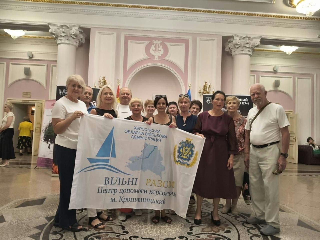 Група херсонських переселенців позує для спільного фото, тримаючи великий офіційний прапор хабу у витонченому класичному фоє Кіровоградського обласного академічного українського музично-драматичного театру імені М. Л. Кропивницького перед початком театральної вистави. На передньому плані учасники зустрічі, організованої координаційним центром «Вільні разом», тримають велике біле полотнище з мапою та гербом області, логотипом у вигляді синього вітрильника, офіційними написами «ХЕРСОНСЬКА ОБЛАСНА ВІЙСЬКОВА АДМІНІСТРАЦІЯ» та «ВІЛЬНІ РАЗОМ Центр допомоги херсонцям м. Кропивницький». Жінки та чоловіки різного віку вбрані у класичний та повсякденний одяг, зокрема у літні світлі футболки, блузки, темні сукні та штани, посміхаючись в об'єктив кадру. Спільний культурний візит для родин ВПО з Херсонщини проходить у розкішній залі театру на тлі високих світлих колон із ліпниною, декоративних арок у рожево-білих тонах із зображенням ліри та масивних дверей, де ліворуч та праворуч помітно рекламні театральні банери-афіші майбутніх вистав.