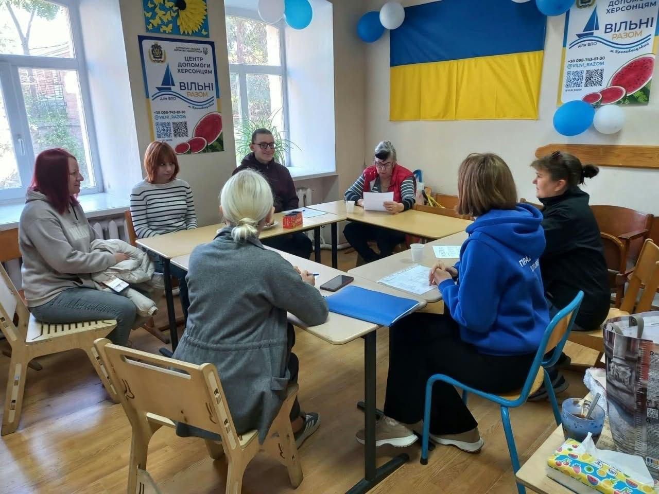 Психологиня благодійного фонду «Право на захист» Ірина Лисенко проводить тренінгову зустріч «Бінго базових потреб» для переселенців із Херсонщини в офісі координаційного центру «Вільні разом» у Кропивницькому. Учасники заходу спілкуються в дружній атмосфері, сидячи навколо великого столу у формі літери «П», заповнюють роздаткові бланки та обговорюють методи відновлення внутрішніх опор. Справа за столом сидить психологиня у фірмовому яскраво-синьому худі організації, а навпроти неї розмістилися відвідувачі у повсякденному одязі та куртках, які уважно слухають наставницю. Психосоціальний практикум для підтримки родин ВПО проходить у світлій кімнаті на тлі Державного прапора України, синьо-білих повітряних кульок під стелею, а також двох ідентичних білих банерів із логотипом «Центр допомоги херсонцям ВІЛЬНІ РАЗОМ», один із яких закріплено на стіні ліворуч біля вікна, а другий розташовано у кутку праворуч.