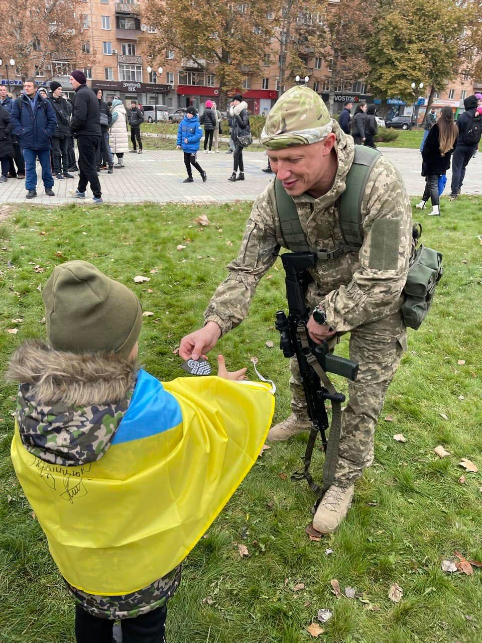 Євгеній Ігнатенко, який саме на момент зйомки цього фото був військовослужбовцем Збройних Сил України, з усмішкою передає невеликий чорно-білий текстильний шеврон дитині, яка загорнута у Державний прапор України на зеленій зоні площі Свободи у звільненому Херсоні. Праворуч у кадрі на повний зріст стоїть Євгеній Олександрович у камуфляжній формі, тактичному розвантажувальному жилеті, світлих берцях та військовій бандані, який злегка нахилився вперед і тримає в лівій руці свій автомат зі спеціальним ременем. На передньому плані ліворуч спиною до об'єктива стоїть маленька дитина в теплій зимовій куртці з камуфляжним капюшоном та в зеленій шапці, плечі якої повністю покриті синьо-жовтим національним стягом із нанесеними маркером написами подяки та малюнком серця. Цей історичний момент щирого спілкування та підтримки зафіксований на території головної площі міста, де на задньому плані вздовж пішохідної доріжки перебуває велика кількість містян, які святкують визволення, а вдалині проглядаються житлові будинки та відомі міські будівлі з вивісками.