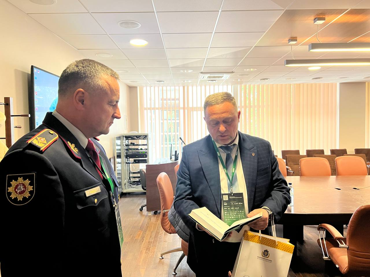 Перший заступник начальника Херсонської ОВА Дмитро Бутрій презентує книгу «Unbreakable Kherson» директору Департаменту пожежної охорони та рятування при МВС Литви генералу внутрішньої служби Ренатасу Пожелі під час офіційної робочої зустрічі. У центрі кадру Дмитро Бутрій у класичному темно-синьому костюмі, білій сорочці з краваткою та з великим брендованим бейджем конференції на шиї тримає в руках відкритий примірник видання, гортаючи сторінки з покроковими рекомендаціями, а в лівій руці тримає світлий паперовий подарунковий пакет із офіційним золотавим логотипом-гербом. Ліворуч боком до об'єктива стоїть генерал Ренатас Пожела у парадній темно-синій формі литовської рятувальної служби з червоними кантами, золотистими погонами, емблемою із зображенням вершника «Вітіса» на правому рукаві та нагородами, який уважно вдивляється в текст книги. Робоча зустріч для координації рятувальних підрозділів проходить у світлому офісному кабінеті, де на задньому плані помітно великий дерев'яний стіл переговорів із коричневими кріслами, телевізійну панель, металеву стійку з технічним обладнанням та велике вікно з вертикальними жалюзі.