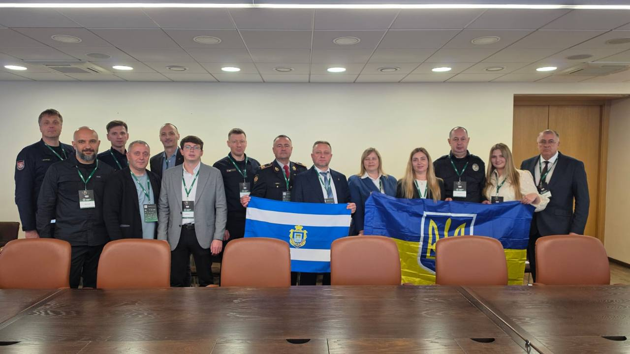 Делегація Херсонщини разом із литовськими партнерами розгортає Державний прапор України та біло-блакитний прапор міста Херсона, позуючи для спільного фото у конференційній залі під час офіційного візиту до Литви. Учасники міжнародної безпекової зустрічі вишикувалися в один щільний ряд за довгим темно-коричневим дерев'яним столом переговорів із помаранчевими шкіряними кріслами. По центру кадру стоїть перший заступник начальника Херсонської ОВА Дмитро Бутрій у класичному темно-синьому костюмі та білій сорочці, який разом із колегами тримає прапор міста із золотим гербом на синьому щиті по центру. Справа від нього представники української делегації розгорнули великий синьо-жовтий національний стяг із золотистим Тризубом, який четвертою праворуч від краю кадру безпосередньо тримає заступниця директора Агенції регіонального розвитку Херсонщини Інна Бутрій у світлій блузці. У лівій частині знімка серед державних службовців Херсонської ОДА та представників громадських організацій другим за рахунком стоїть Юліан Лучко у чорному світшоті з акредитаційним бейджем на шиї. Поруч із Дмитром Бутрієм ліворуч стоять керівники рятувальних та правоохоронних служб у темному форменому одязі з нашивками, зокрема директор Департаменту пожежної охорони та рятування при МВС Литви генерал Ренатас Пожела у парадній формі з нагородами, а з правого краю кадру розмістився начальник Дарʼївської сільської військової адміністрації Анатолій Дідур у чорному костюмі, білій сорочці та краватці, замикаючи спільний патріотичний знімок у світлому сучасному кабінеті під лінійними стельовими світильниками.
