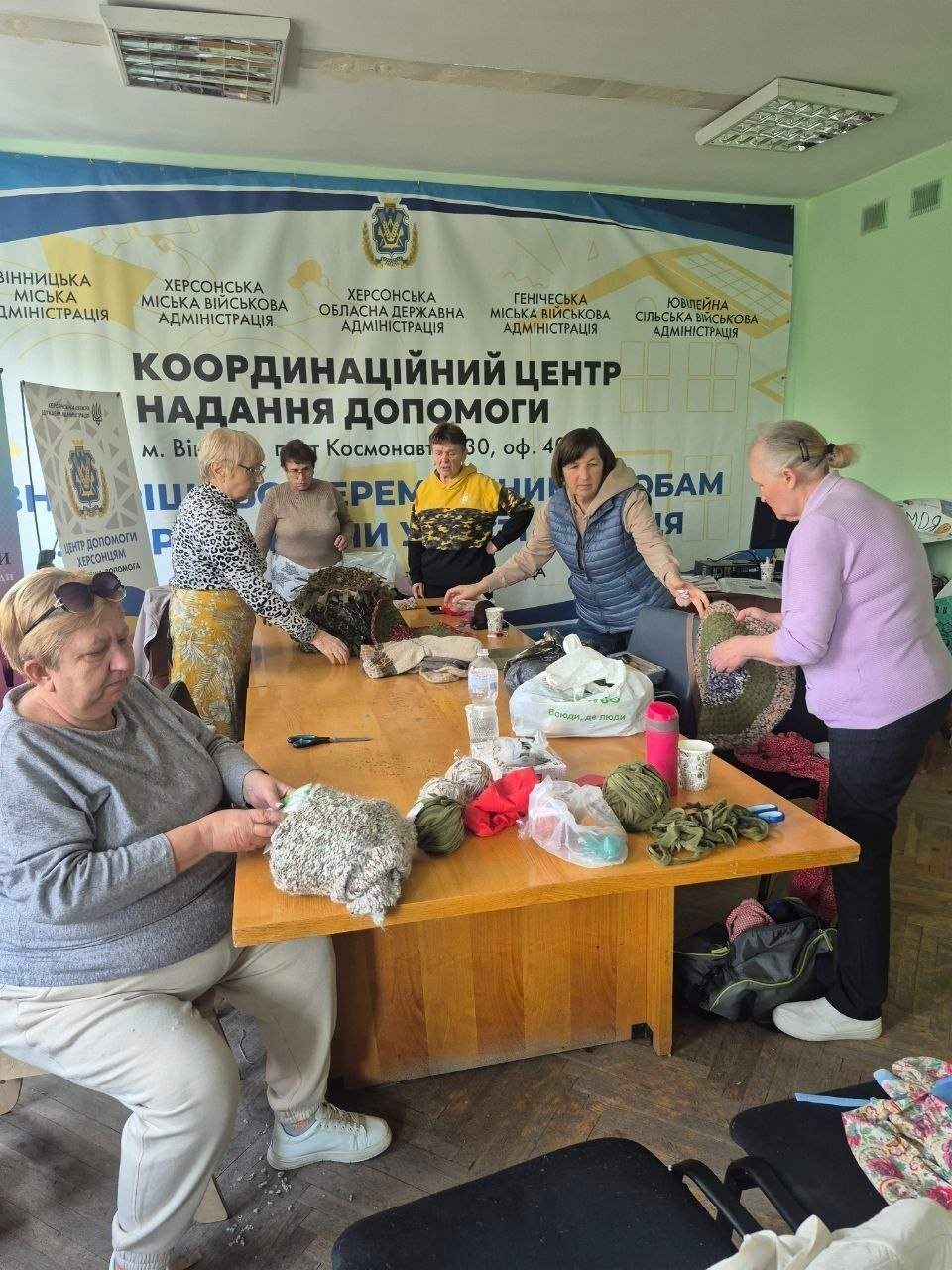 Переселенки з Херсонщини зосереджено плетуть захисні нашоломники та в'яжуть теплі шкарпетки для військовослужбовців Збройних Сил України, зібравшись за великим спільним столом у вінницькому координаційному центрі «Вільні разом». Жінки старшого та середнього віку в повсякденному теплому одязі, кофтах і жилетках розклали на довгому дерев'яному столі мотки зелених, коричневих та сірих ниток, ножиці, заготовки маскувальних сіток і пакунки з матеріалами. Вони активно працюють руками, створюючи елементи захисного спорядження для наших захисників на передовій. Творча волонтерська зустріч для системної підтримки армії та психосоціальної взаємодії проходить у просторому світлому кабінеті хабу, де на задньому плані стіну повністю закриває великий офіційний біло-зелений банер із гербом та текстом «КООРДИНАЦІЙНИЙ ЦЕНТР НАДАННЯ ДОПОМОГИ м. Вінниця», а також офіційними назвами Херсонської ОДА, Херсонської, Генічеської та Ювілейної міських і сільських військових адміністрацій.